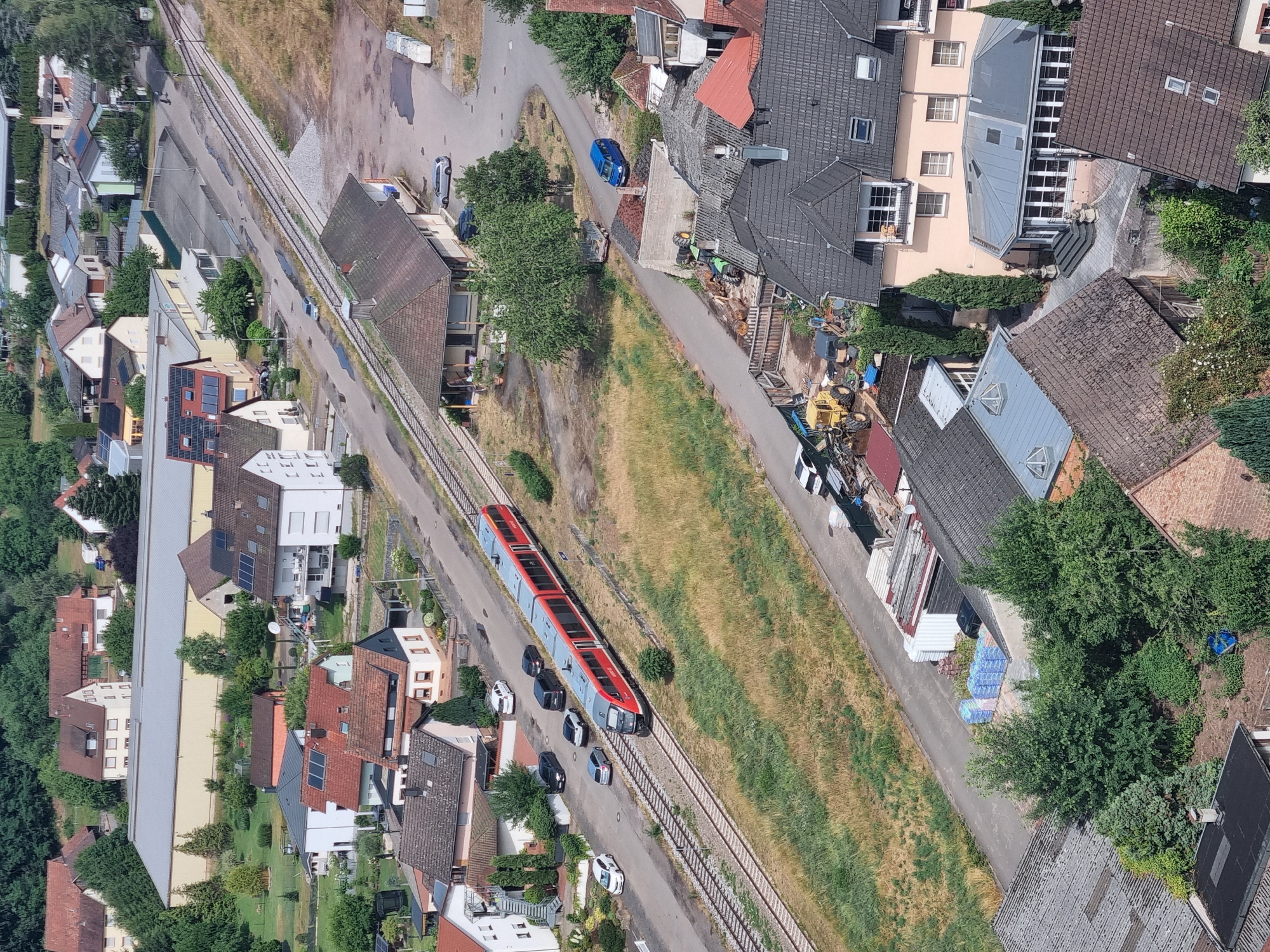 This aerial view depicts a train, likely a regional passenger train, traveling on a railway line that runs through a suburban or semi-rural area. The train is red and silver and appears to be moving from left to right in the frame. Several cars are parked along a road that runs parallel to the tracks. The scene is populated with residential buildings, some with red tile roofs and others with modern, flat roofs. There are also some commercial or industrial buildings with large, light-colored roofs. Green trees and vegetation are interspersed throughout the landscape, with dry, grassy areas suggesting a warm, possibly dry season. The time of day appears to be daytime, with clear lighting casting shadows. The overall impression is one of a quiet, functional community with transportation infrastructure integrated into the residential areas. No visible text is present in the image. The context suggests this location is Dahn, Germany.