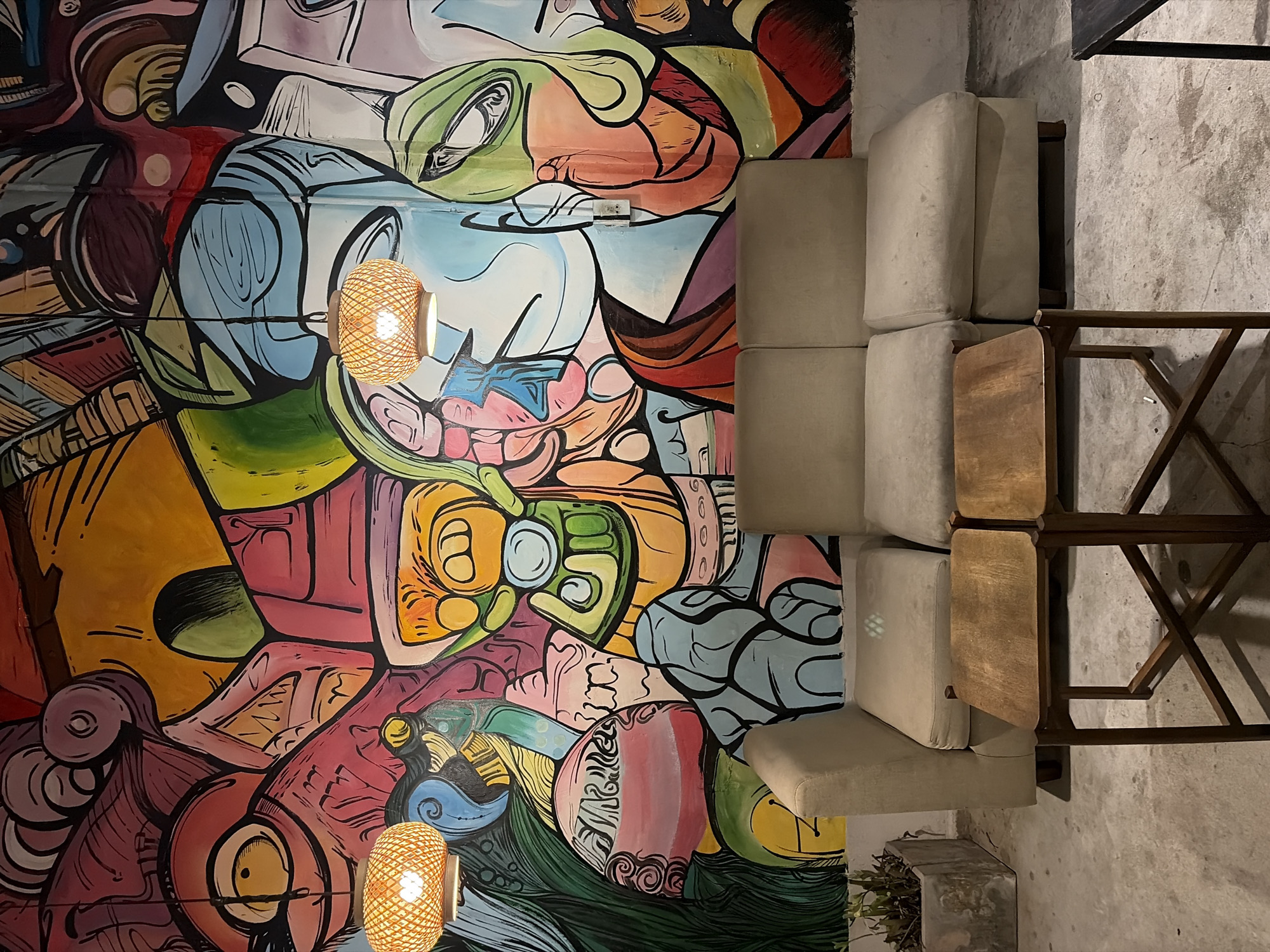 The image depicts an indoor seating area, likely a cafe or lounge, with a prominently featured vibrant mural covering the entire back wall. The mural is an abstract, colorful composition with bold lines and diverse shapes, featuring elements that suggest stylized faces and organic forms in shades of pink, yellow, blue, green, and orange. Two woven bamboo pendant lamps hang from the ceiling, illuminating the space below. In front of the mural, a light beige modular sofa with multiple cushions sits on a concrete floor. Two small, square wooden coffee tables with dark wooden legs are positioned in front of the sofa. To the left of the sofa, a rectangular concrete planter contains a few dry, leafy plants. The overall ambiance suggests a contemporary and artistic urban setting. The scene is devoid of any people.