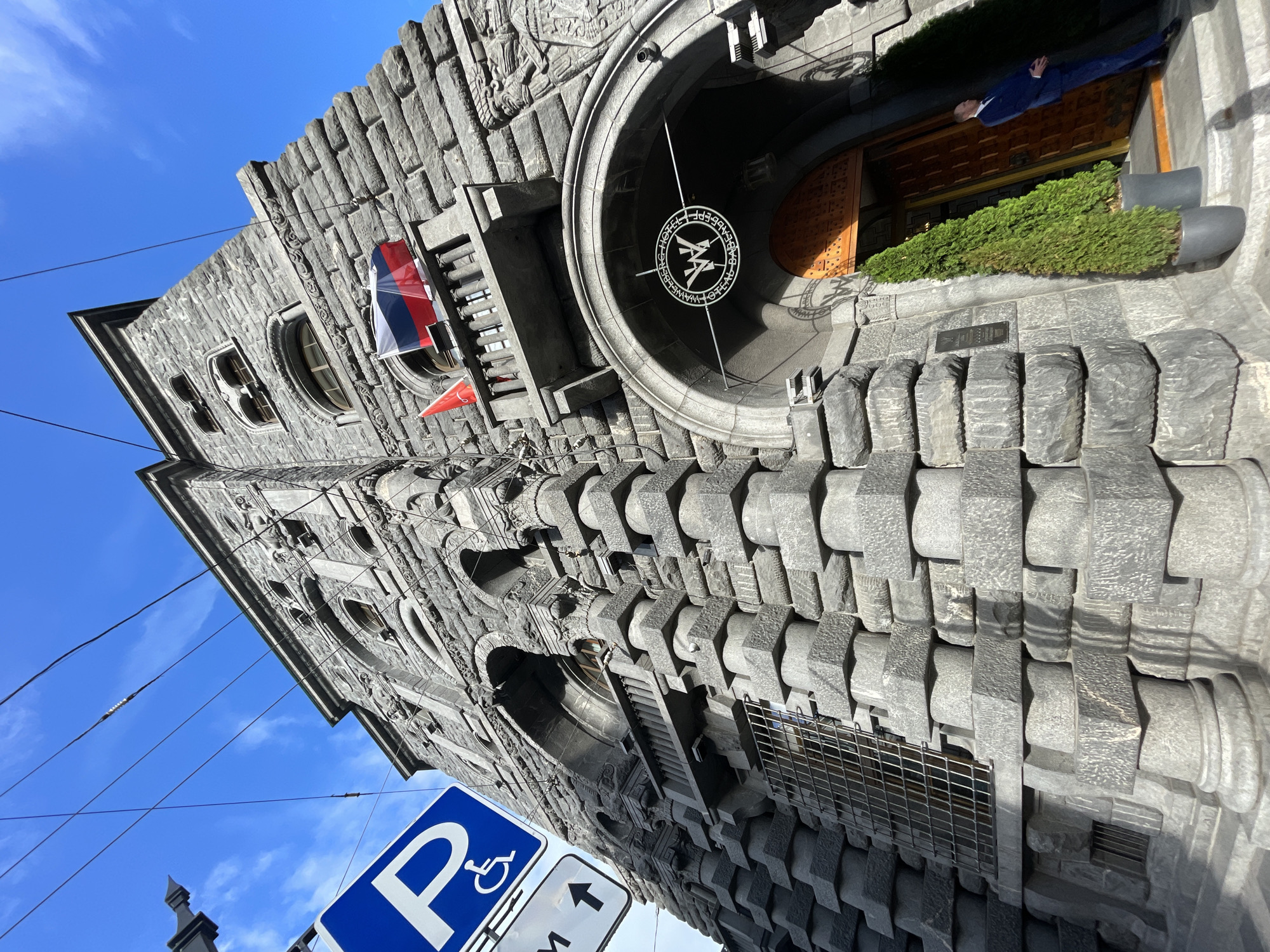 The image shows the Wawelberg Hotel in Saint Petersburg, Russia. It is a historic building with a distinctive granite facade featuring intricate carvings and architectural details. The building is tall and imposing, with a prominent arched entrance. A Russian flag is displayed on a balcony on the upper floors. A sign with the hotel's logo is visible above the entrance. A man in a suit stands near the entrance, and two potted evergreen trees flank the doorway. A blue parking sign for disabled access is visible in the upper left corner of the image, along with overhead electrical wires. The sky is clear and blue with some scattered clouds.
