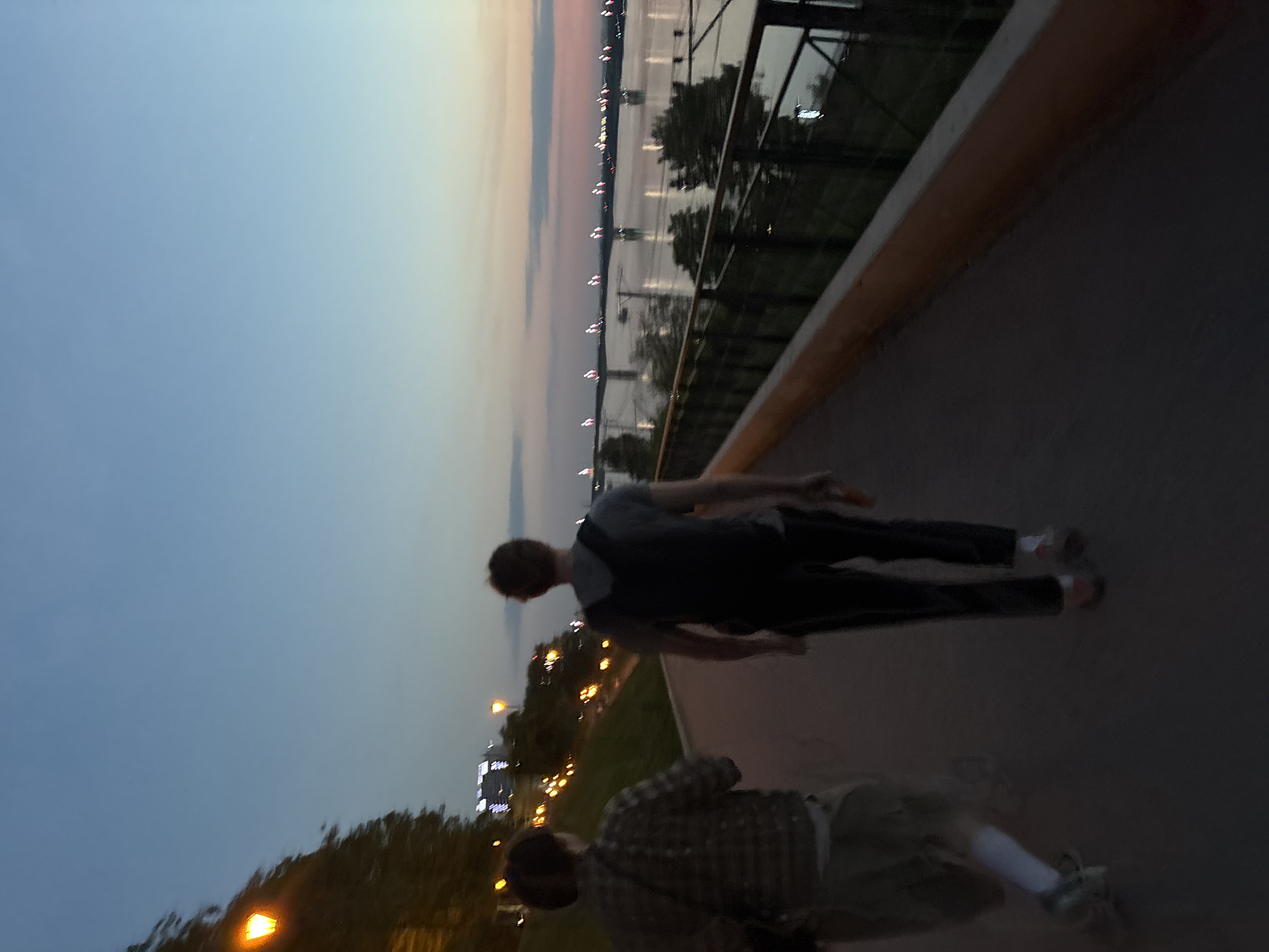 Two male figures are captured on a path beside a fence, likely near the Kama River in Perm, Russia, during the twilight hours. The riverbank is illuminated by city lights across the water. One individual is wearing a dark t-shirt and dark pants. The other person wears a checkered shirt and light-colored shorts. Both appear to be walking along the path.
