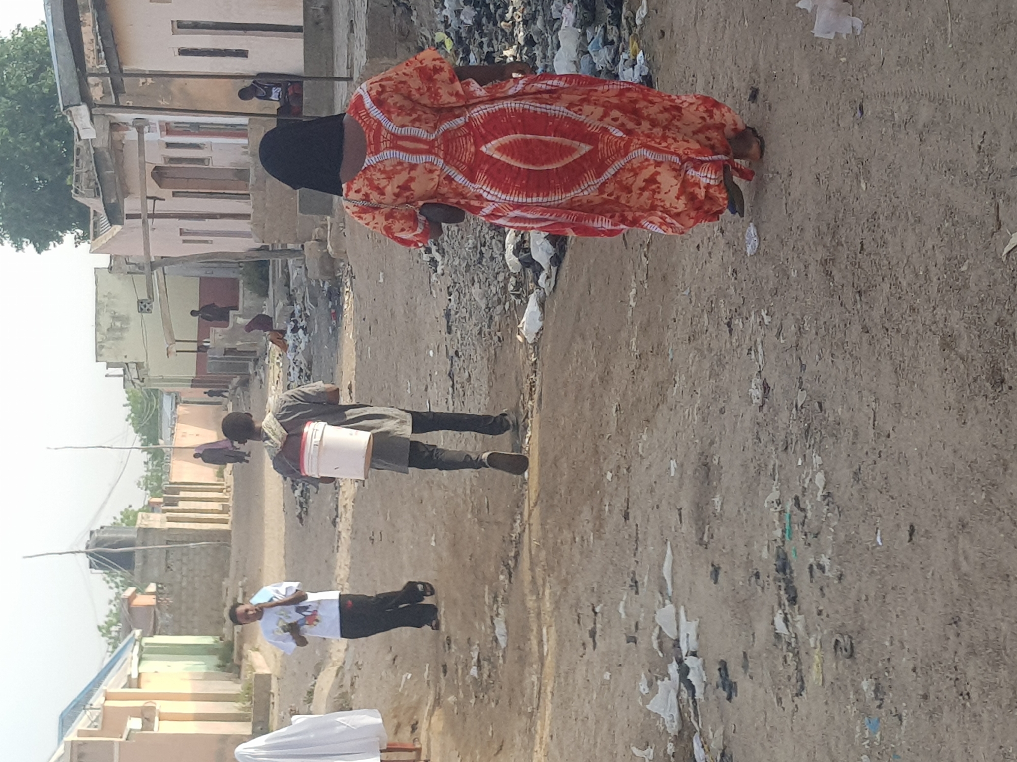 The image shows a street scene in what appears to be the Dutsinma B area of Nigeria. The road is unpaved, and littered with trash. Buildings line both sides of the street, showing signs of wear and limited infrastructure. Several people are visible: a woman in a patterned orange dress and a dark head covering walks toward the center of the image, apparently near a pile of trash. A man walks down the street carrying a white bucket. A young man walks down the street in a white t-shirt with dark pants. Other people are seen further down the street.