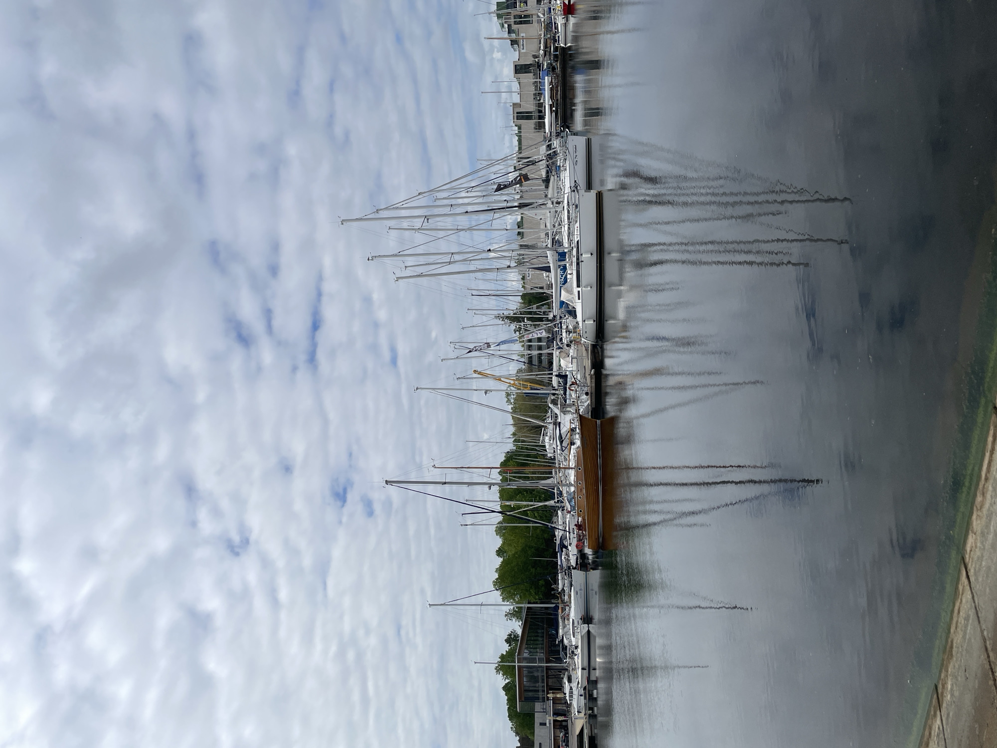The image shows a harbor in Tallinn, Estonia, filled with sailboats and yachts. The water reflects the masts and hulls of the boats, as well as the cloudy sky above. There are some buildings and trees visible in the background along the waterfront.