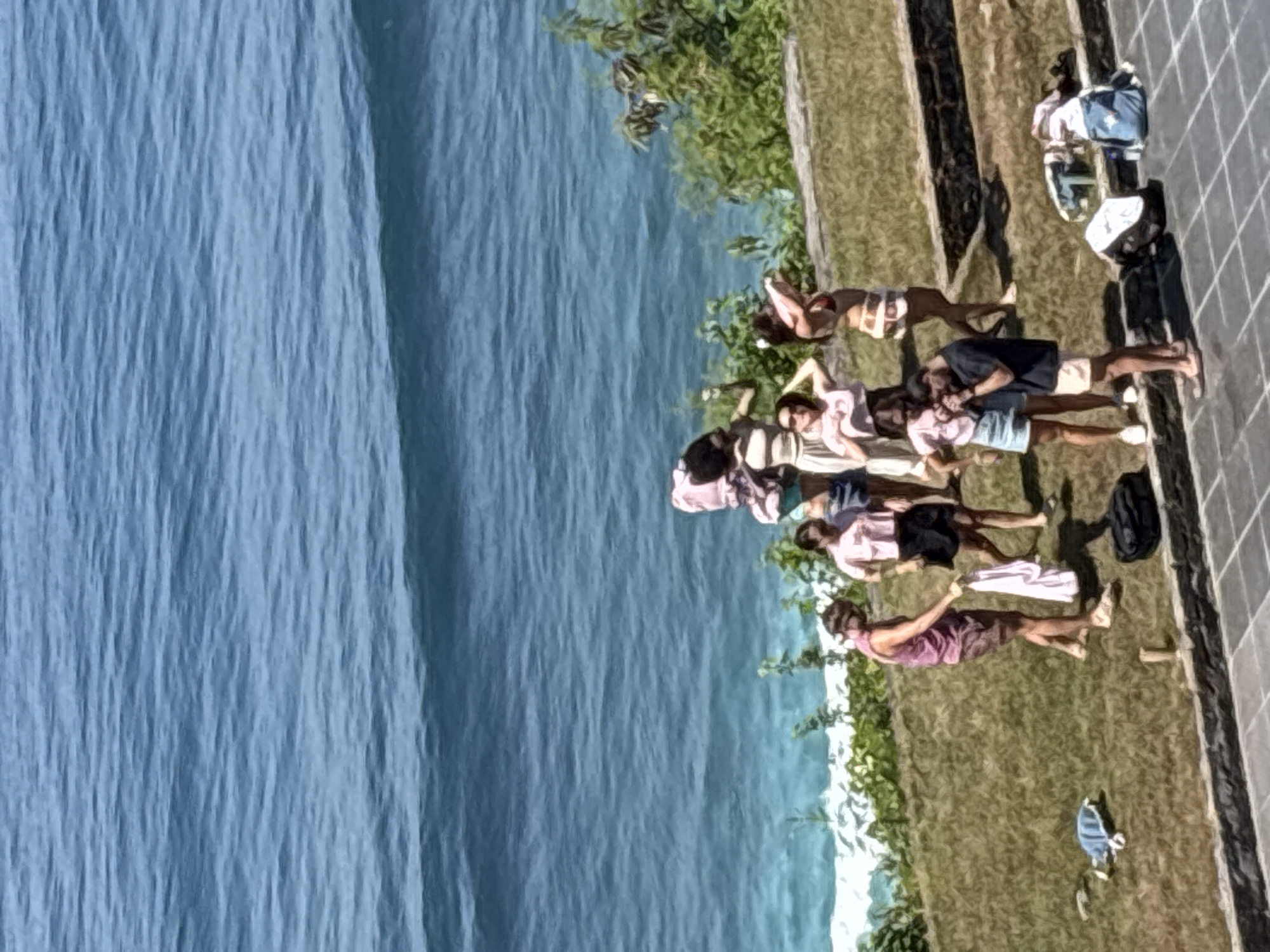 The image shows a group of people lounging near the ocean. The location is likely in Kuta Selatan, Indonesia, as it features a shoreline with blue water on the left and a grassy area on the right. Several individuals are lying down on the grass. Some are oriented toward the ocean while others are facing away from it. They are casually dressed in shorts, shirts, and swimwear. A few objects, including a black backpack, a white cloth, and a blue bag, are scattered nearby. A paved area with some people on it is along the right edge of the image.