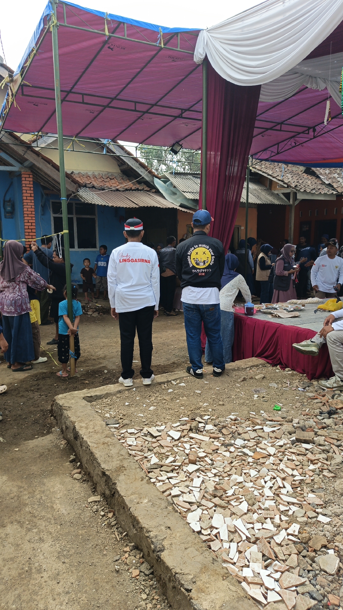 The image depicts an outdoor gathering, possibly a community event, in Linggasirna, Indonesia. In the foreground, two individuals stand facing away from the camera, one wearing a white long-sleeved shirt with red and white headwear and the text "LINGGASIRNA" in red, the other in a black jacket with yellow text and a blue cap. A young child in a blue shirt and dark patterned shorts stands to the left, holding a wooden pole.
The scene is set under a large, pink and blue tent structure, supported by poles. Burgundy drapery hangs from the tent's edge. In the background, several other people are visible, some standing and others gathered around a table covered with a red cloth, where food and drinks are laid out. Buildings with tiled roofs are present behind the gathering. The ground is a mixture of dirt and scattered debris, including broken tiles and bricks. The overall atmosphere suggests a casual, communal event taking place in a residential or semi-rural area.