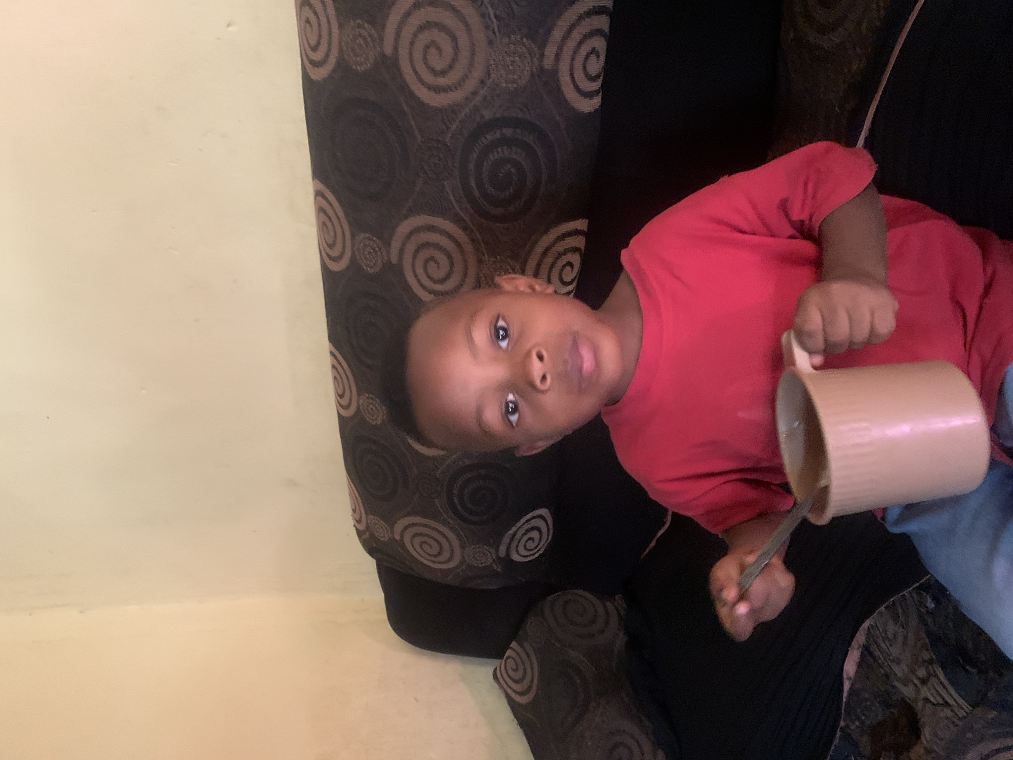 A young child, likely a boy, is depicted sitting or lying on a sofa, looking directly at the camera. He wears a short-sleeved red t-shirt and light-colored pants. In his right hand, he holds a light brown mug with a silver spoon inside it, suggesting he is either drinking or stirring a beverage.
The child is positioned on a dark-colored sofa featuring a distinctive pattern of concentric brown and black spirals or swirls. The background to the left of the child is a plain, light beige wall. The setting appears to be an indoor space, possibly a living room. The lighting is soft and even, indicating either diffused daylight or artificial indoor lighting. The child's expression is neutral and observant. There is no visible text in the image. The scene captures a quiet, everyday moment.