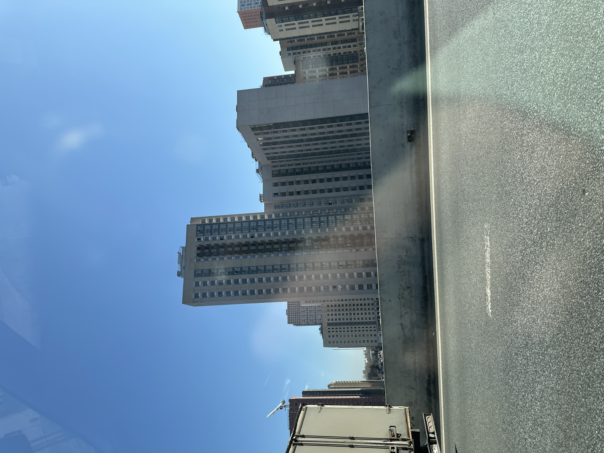 The image shows a view from a vehicle driving on a highway in Moscow, Russia. Buildings line the roadside, including high-rise residential structures. The road surface is asphalt, with a painted white lane marker. The sky is blue with some clouds. A truck or trailer is partially visible in the foreground.