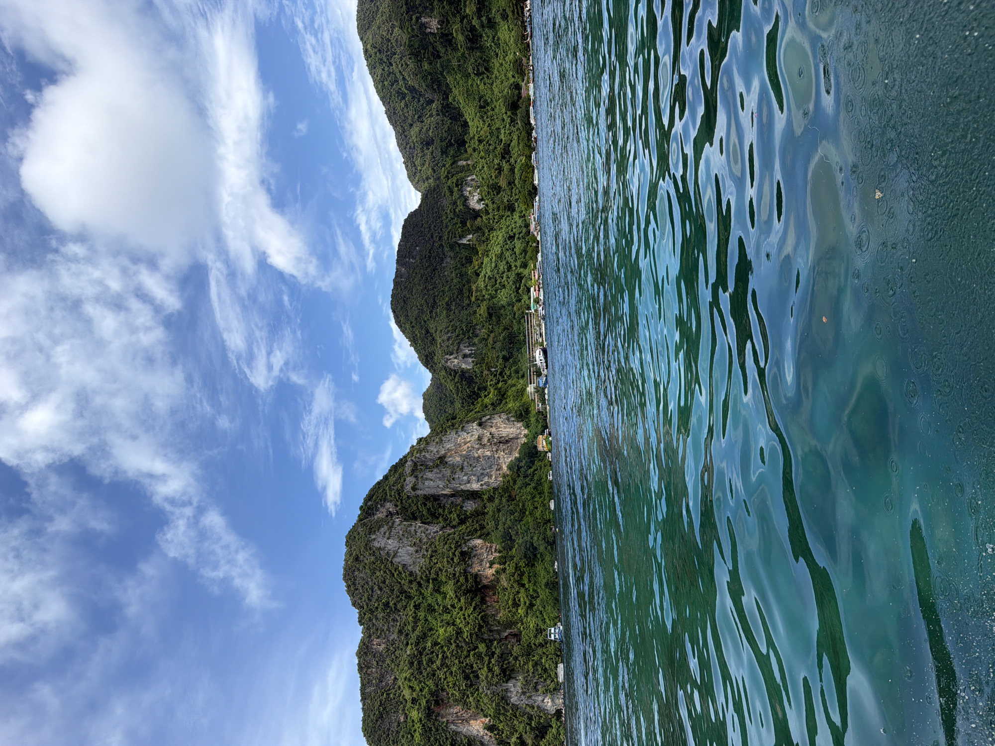 This image captures a scenic view of Ao Nang, Thailand, from the perspective of a body of water. In the foreground, the turquoise ocean is calm with gentle ripples, reflecting the light and sky. Several small boats are visible scattered across the water's surface. In the middle ground, a coastline is lined with lush, green, tree-covered mountains. Some structures, possibly buildings or resorts, are nestled along the base of the mountains, hinting at human habitation. The sky above is a vibrant blue, dotted with fluffy white clouds. The overall impression is a serene tropical landscape.