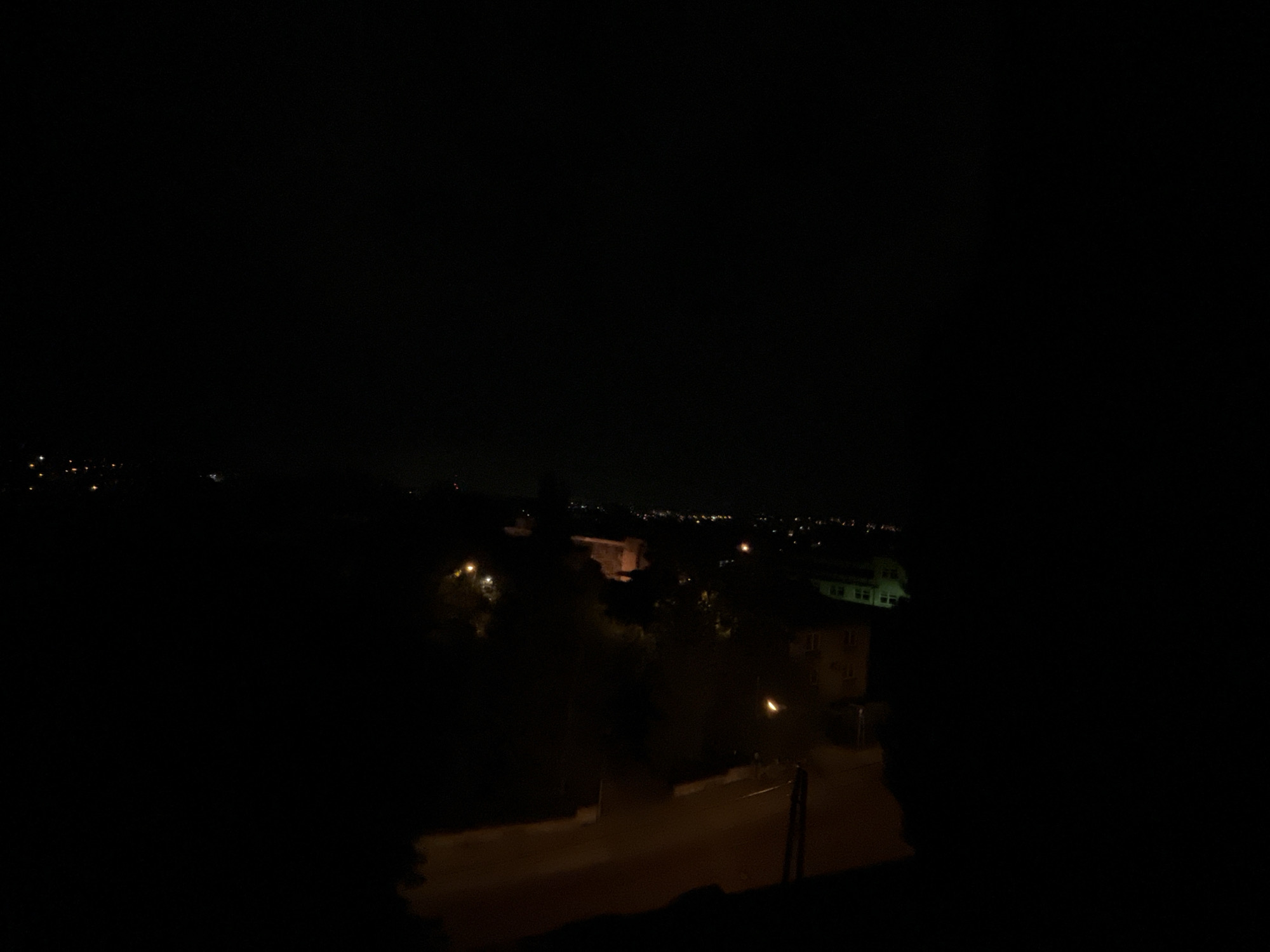 A nighttime scene depicts a residential area in Unknown. The composition includes a road with street lights, houses with illuminated windows, and trees. Distant city lights are visible in the background. The scene appears quiet and still.