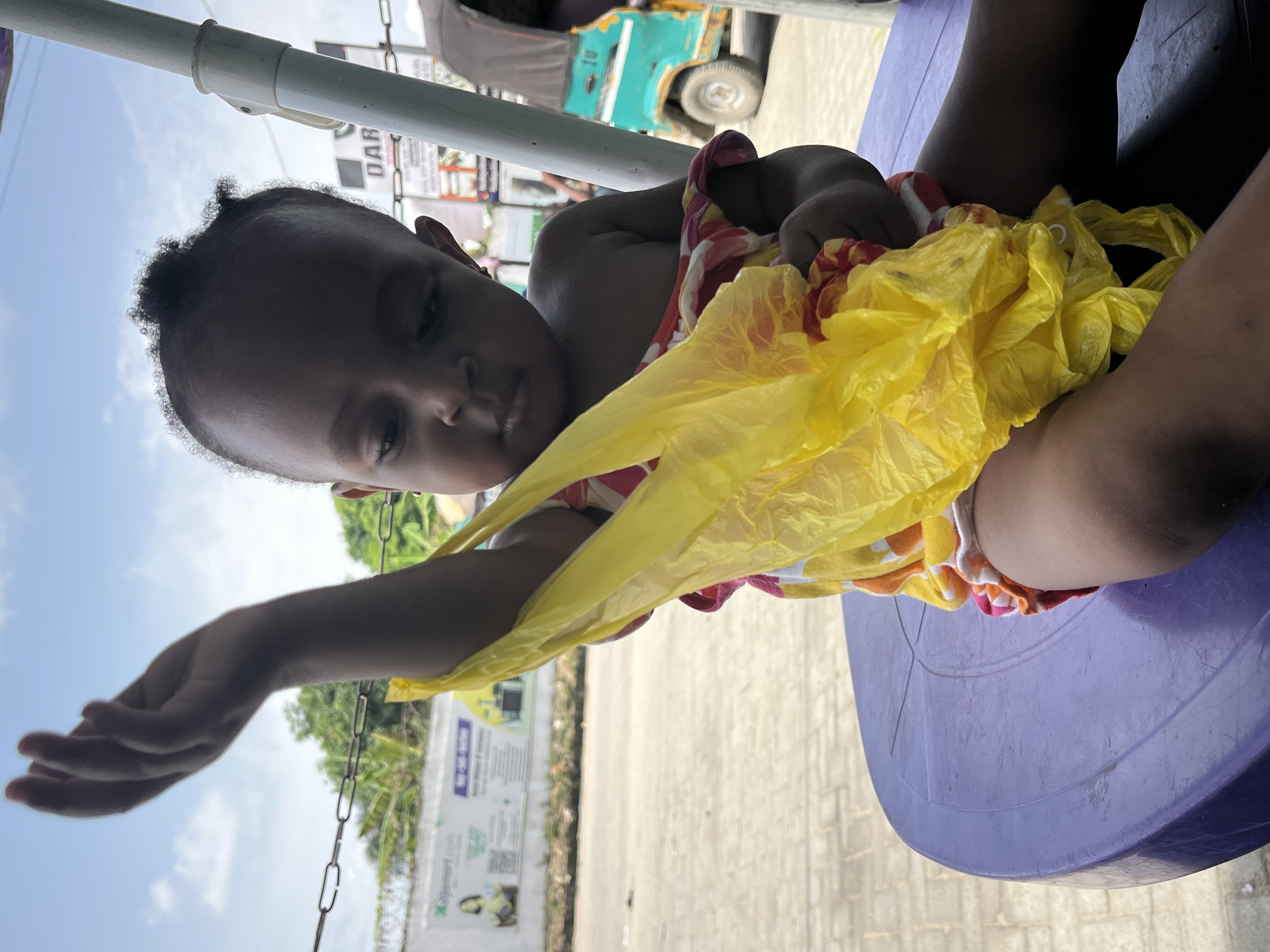 A child sits on a purple plastic stool. The child holds a bunch of yellow plastic bags. The location is Lekki, Nigeria, as evidenced by a sign in the background that reads "Mart.com.ng" and other details of the physical environment. A tuk-tuk is visible in the background.