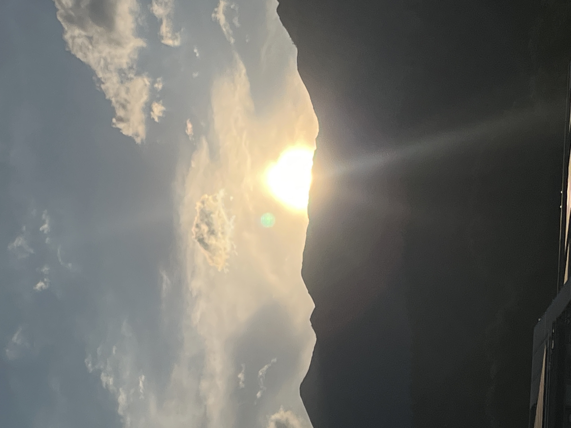 This media file depicts a landscape scene, likely taken in Garmisch-Partenkirchen, Germany. The dominant subjects are a mountain range silhouetted against a sky filled with clouds. The sun is brightly visible, partially obscured by the mountain, casting a warm glow and creating lens flare, suggesting it is either sunrise or sunset. The sky transitions from a bright, sunlit area with scattered clouds to a darker, more overcast portion. The mountain's edge is sharply defined, contrasting with the softer forms of the clouds. The overall mood conveyed is serene and awe-inspiring, highlighting the natural beauty of the mountainous terrain at a dramatic time of day. There are no visible people or objects in the immediate foreground, and no text is present in the image.
