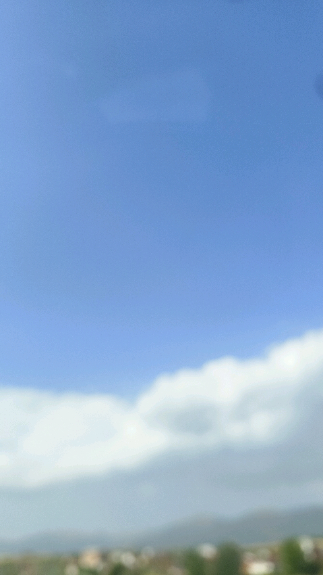 The image is a blurry, vertical shot of a bright blue sky with a band of white clouds across the lower portion, above a blurred landscape of rolling hills and what appears to be a settlement or town. The lower edge of the image shows a suggestion of trees and buildings. The sky is clear and the light suggests daytime. There are no discernible people, subjects, or objects, and no actions or interactions are depicted due to the significant blurriness. The setting appears to be outdoors, likely a natural or semi-natural environment with a distant view of inhabited land. It is not possible to identify any specific location within Skopje, North Macedonia, due to the lack of identifiable landmarks or details.