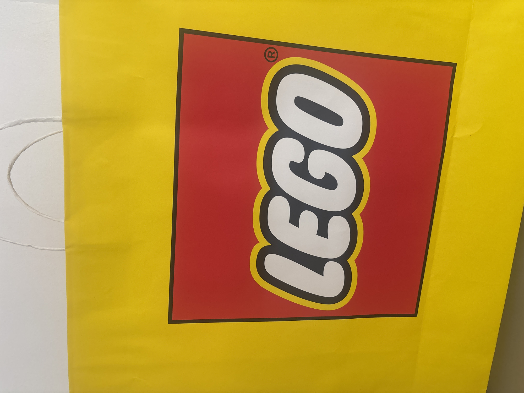 A close-up shot depicts a yellow LEGO shopping bag. The iconic LEGO logo, consisting of white block letters outlined in yellow on a red background, is prominently displayed. The bag appears to be against a white wall. The handle of the bag is visible on the left side. The location is not specified in the image, so providing the information about the city of San Pablo del Monte, Mexico is not possible.