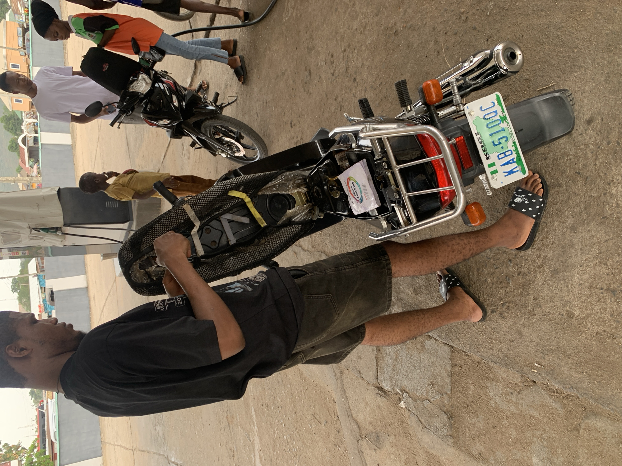 A street scene in Lokoja, Nigeria, shows a man in a black t-shirt and dark shorts standing next to a motorcycle. The motorcycle has a license plate KAB-510UC. Other individuals are nearby: a man in a white t-shirt, and others. The setting appears to be a public area, possibly near a gas station.