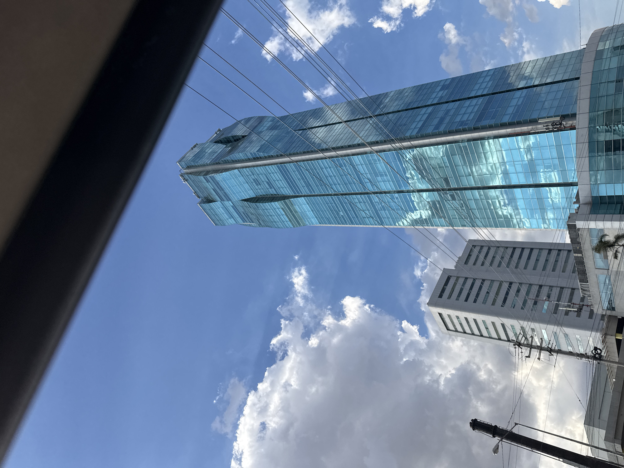 The image shows a view looking up at two buildings in what is likely San Bernardino Tlaxcalancingo, Mexico, given the prompt's location requirement.
The primary building is a tall skyscraper with a blue glass facade reflecting the sky. Its design appears modern and stepped. Overhead electrical or communication wires are visible against the sky and building.
To the right of the skyscraper, a shorter, block-shaped building with a white or light grey exterior is visible. This building has a repetitive pattern of windows arranged in vertical columns.
The sky is blue with scattered white cumulus clouds. The perspective is angled upwards, indicating the photograph was taken from street level or from within a vehicle.
A portion of a dark, possibly black, object is present in the left of the frame, potentially a part of the vehicle's interior from where the photo was taken. A utility pole and other urban infrastructure are partially visible in the bottom right corner. There are no discernible people in the frame. The environment appears to be a city with a modern architecture.