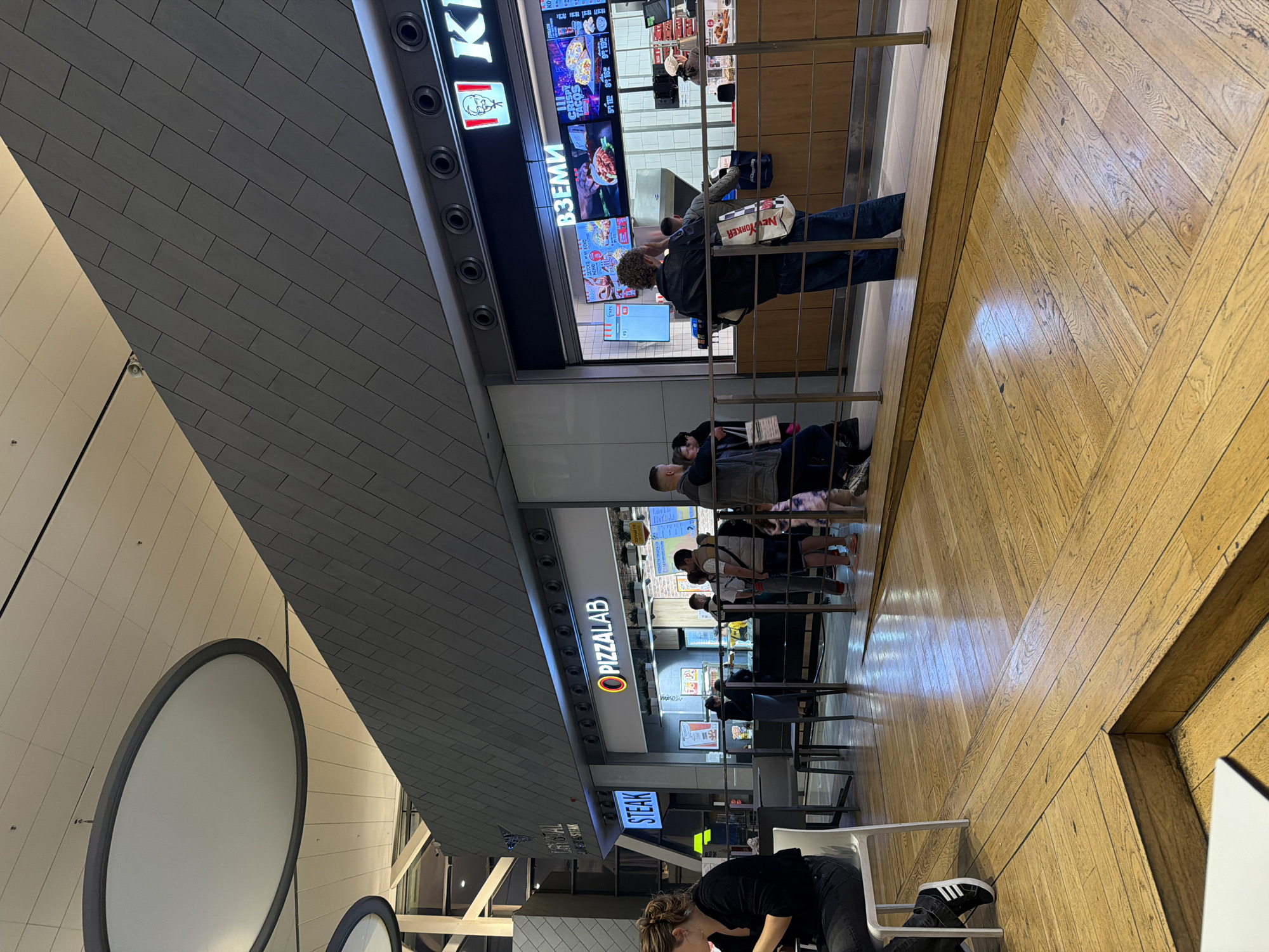 This image depicts an indoor food court or commercial area, with a wooden floor in the foreground and various food establishments in the background. The floor has a distinctive herringbone or chevron pattern of light-toned wooden planks.
In the bottom right, a person with brown hair, wearing a black top and dark pants with white-striped Adidas sneakers, is seated at a white table, looking down. The table is partially visible.
A metal railing separates the foreground seating area from the bustling food stalls in the mid-ground. A queue of people is visible, waiting to order food. On the left side of the background, a KFC outlet is prominent, identified by its red and white logo and name. Several digital menu screens display food items, including "CRISPY TACOS" and the Cyrillic word "ВЗЕМИ" (meaning "TAKE" or "GET"), along with prices. A person in a dark jacket and jeans, holding a white shopping bag emblazoned with "NEW YORKER," stands in the KFC queue. Further along the line, other individuals are waiting.
Next to KFC, another food stall sign reads "PIZZALAB" with an orange circular logo. Beyond PIZZALAB, another illuminated sign clearly shows the word "STEAK." The ceiling above the food stalls is constructed with gray rectangular tiles, creating a geometric pattern. To the far left, a light-colored wall with large, circular, dark-rimmed decorative elements suggests modern architecture.
The scene appears to be a typical moment in a busy public space, likely a mall or airport terminal, with people either waiting for food or seated. The lighting is bright and artificial, consistent with an indoor setting.
