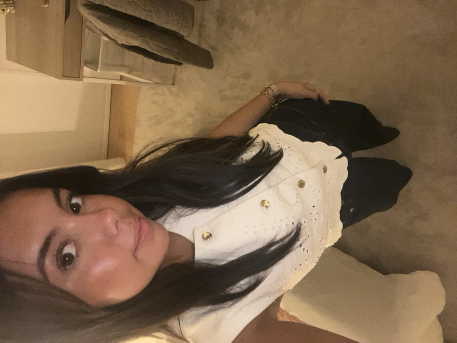An indoor, vertical, full-body image portrays an adult female from a high-angle perspective, appearing as a self-portrait. The subject possesses long dark hair and wears a white, sleeveless top featuring eyelet embroidery and four visible gold-toned buttons, paired with dark trousers. Multiple bracelets adorn her left wrist, and a ring is visible on her left ring finger. She looks directly at the camera with a neutral to slight smile. The environment consists of a room with a light-colored carpet covering most of the floor, transitioning to light wooden flooring in the upper right. A light-colored wooden desk with a single drawer and a white metal frame is positioned in the background to the right. A square white bin is located beneath the desk. A quilted, olive-toned garment hangs draped over an object behind the desk. A light-colored upholstered item or cushion is partially visible on the left. Illumination is soft and warm, indicative of artificial indoor lighting. The precise location within Lordelo do Ouro e Massarelos, Portugal, is not discernible from the visual content.