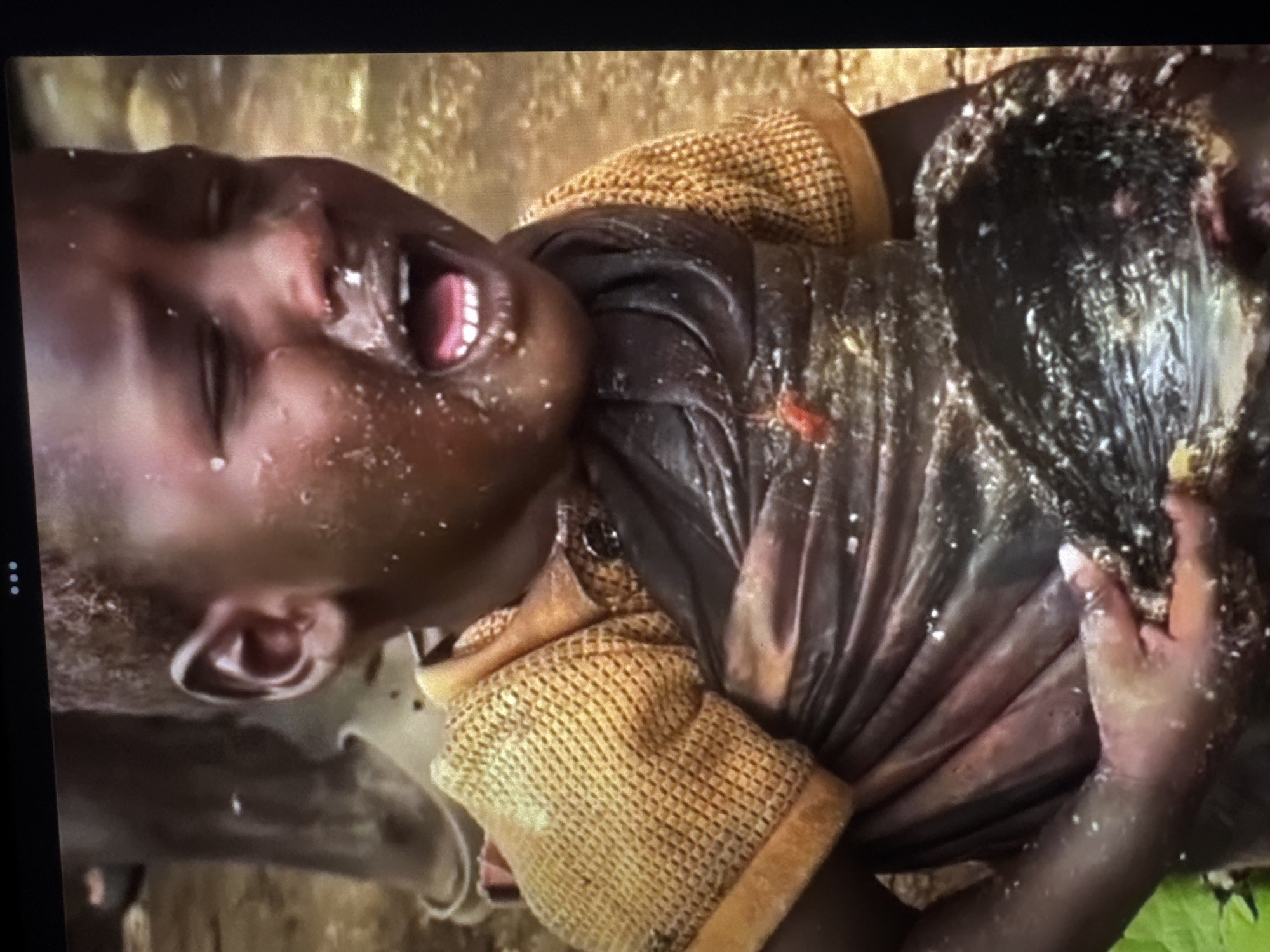 A close-up, over-the-shoulder shot shows a child with their mouth open and eyes closed, appearing distressed. The child is wearing a textured, mustard-colored garment on their upper body and a darker, possibly wet, fabric draped over them. Their skin is dark and appears to have some white specks on it, possibly from water or dirt. The child's right arm is visible, holding a dark, textured object that looks like a bowl or container filled with a dark, viscous substance. A small red speck is visible on the fabric near the child's chest. The background is out of focus but appears to be a natural, outdoor setting with muted, earthy tones. The lighting suggests it could be daytime. The overall impression is one of struggle or hardship. No text is visible in the image.
