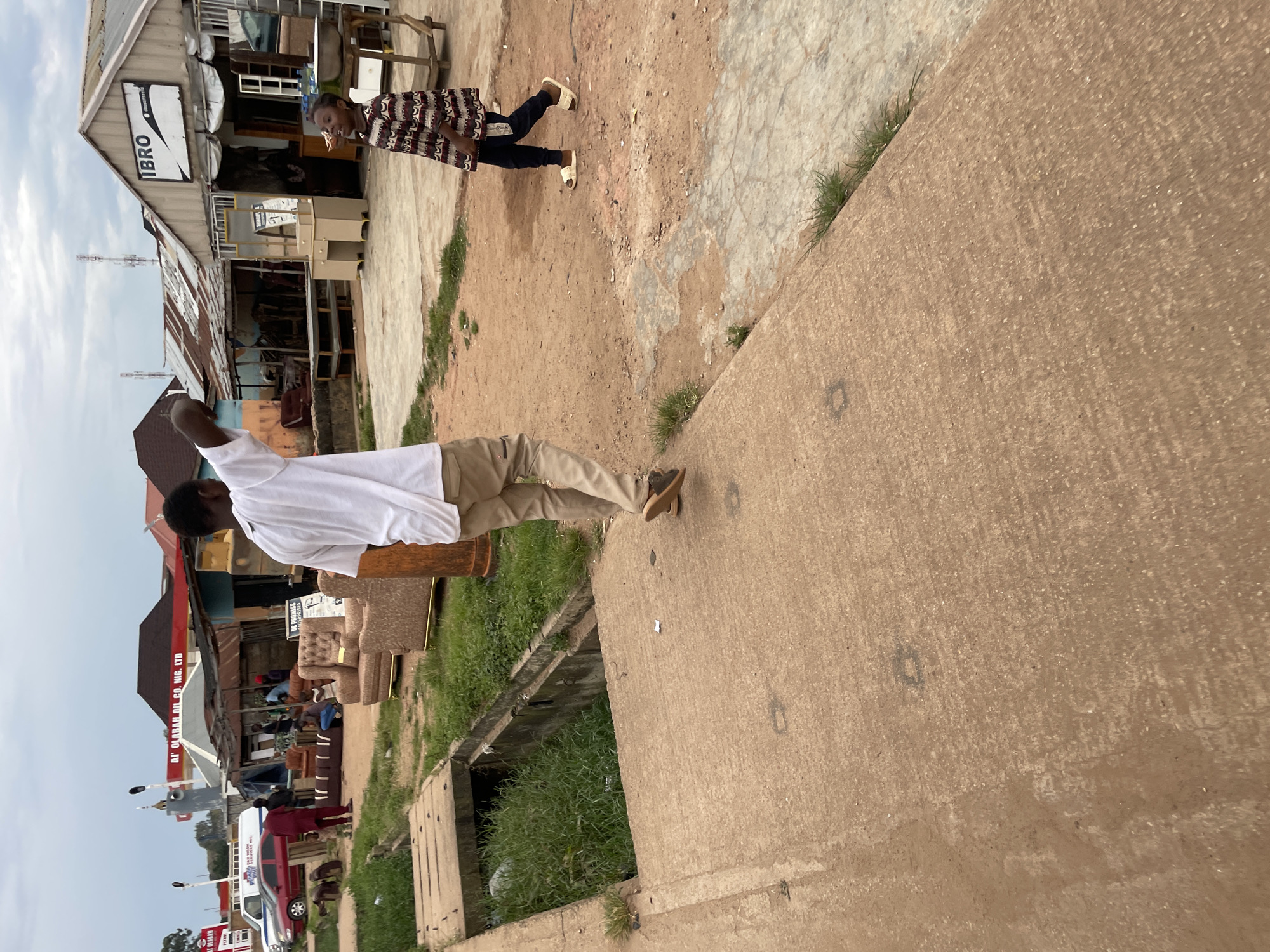 In Barakin Lerawi, Nigeria, a street scene unfolds with a focus on a commercial area featuring various shops and outdoor displays. In the foreground, a young Black male, wearing a white short-sleeved shirt and tan trousers, is walking away from the camera. He is in the process of raising his left arm and pointing, possibly at something off-camera. To his right, a young Black female, dressed in a patterned long-sleeved dress and dark trousers, is looking upwards and slightly to her left, her mouth open. In the background, several structures with corrugated metal roofs house businesses. One prominent sign reads "IBRO" with a phone number. Furniture, including couches and a vanity with a mirror, is displayed outside the shops, suggesting a furniture market or sales area. A red car is parked to the left, and a white van is also visible. A gas station with red and white signage is in the distance. A concrete drainage ditch with overgrown grass runs alongside a paved walkway in the left portion of the image. The overall environment appears to be a bustling, open-air market or commercial strip under an overcast sky.