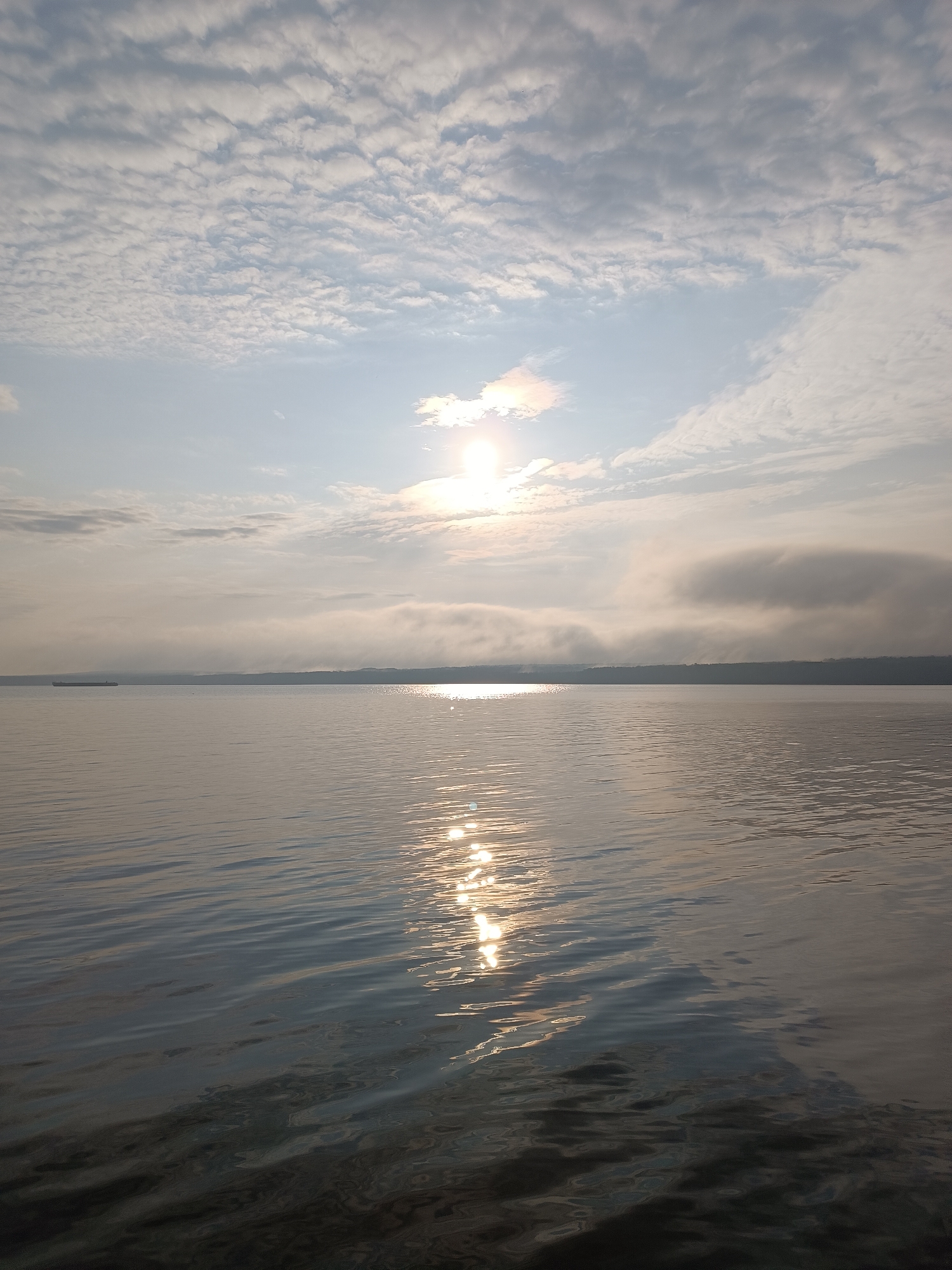 The image captures a serene, expansive view of the Kama River at dawn or dusk in Chaikovsky, Russia. The sun, partially obscured by a thin layer of clouds, casts a warm, shimmering reflection on the water's surface. The sky is filled with a textured pattern of cumulus clouds, with patches of clear blue sky peeking through. A distant, dark tree line marks the horizon. In the middle ground, a long, flat-bottomed barge is visible on the water, suggesting industrial or cargo activity. The water itself is calm, with gentle ripples disturbing the smooth surface, further reflecting the light of the sky. The overall atmosphere is one of tranquility and natural beauty.