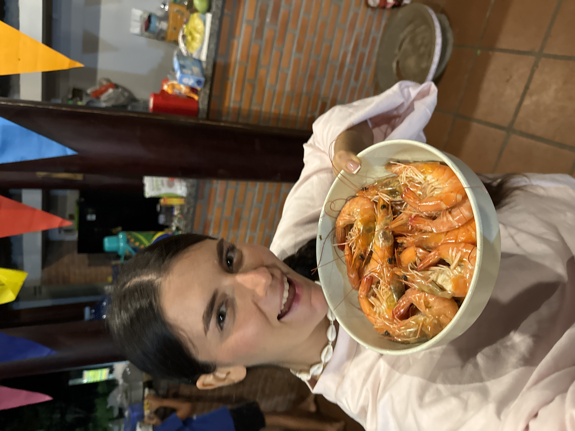 A cheerful woman is depicted holding a white bowl filled with numerous cooked prawns. She has dark hair, parted in the middle, and is smiling broadly directly at the camera, conveying happiness. She wears a light-colored top, possibly pink, and a necklace adorned with white seashells. Her fingernails are manicured with a French tip design.
The scene appears to be an evening or nighttime gathering, indicated by the artificial lighting and the generally dark background. Festive triangular flags in yellow, blue, and red are strung across the upper left side of the frame, suggesting a celebratory atmosphere or party. In the background, a section of a brick wall is visible on the right, and a dark wooden shelf holds various objects, including a red cylindrical item and some small boxes. The floor beneath appears to be composed of terracotta-colored tiles.
The activity centers around a social event where food, specifically seafood, is being enjoyed. The woman's expression and the decorations indicate a relaxed and joyful occasion. Given the provided context of Mũi Né, Vietnam, this setting is consistent with a casual gathering in a coastal region known for its seafood. No visible text is present in the image.