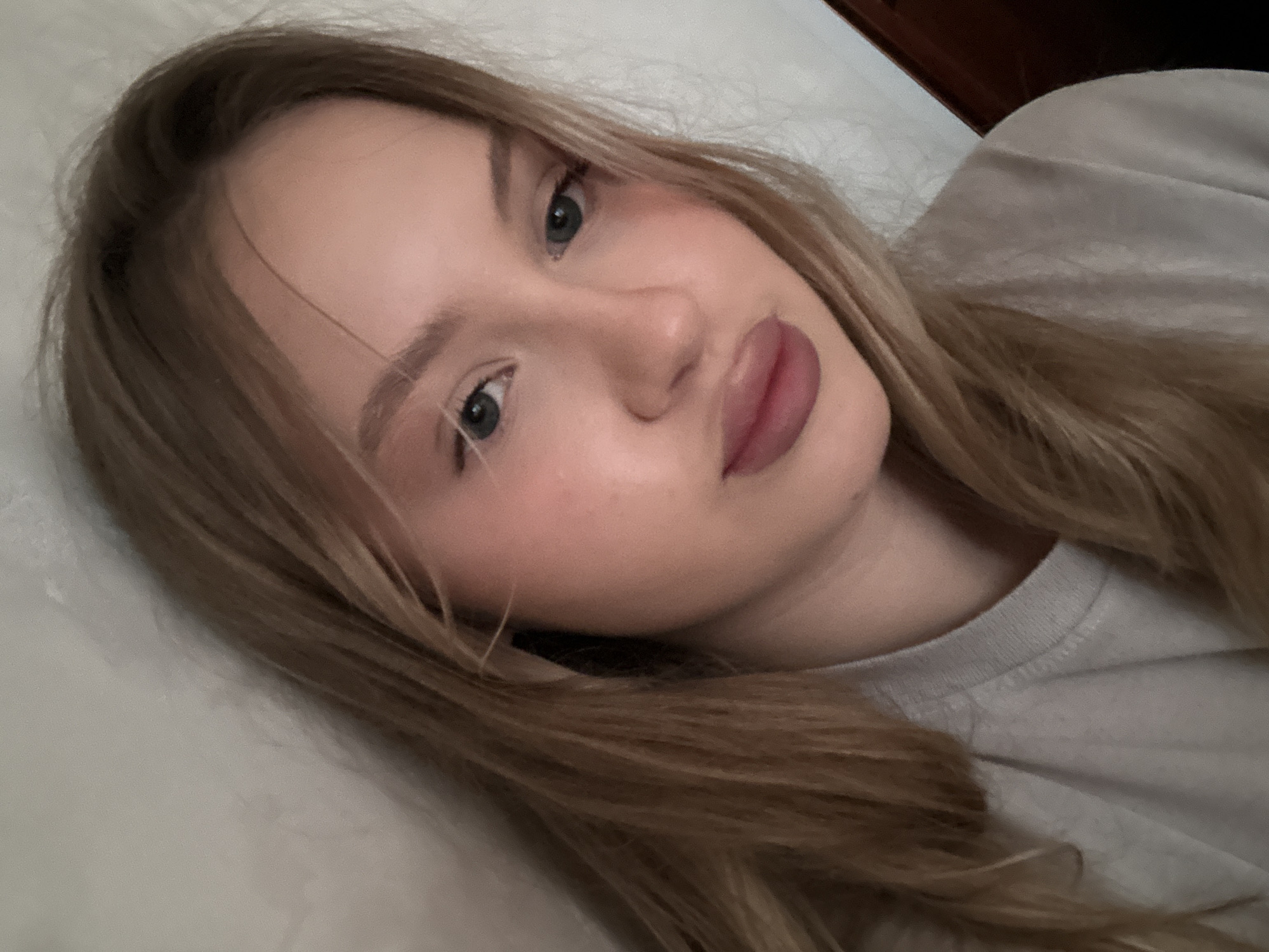A young Caucasian female with blonde hair and fair skin is shown in a close-up, head and shoulders shot. She is wearing a light gray t-shirt. Her head is tilted slightly to her left, and she is looking directly at the camera. Her lips are slightly parted and appear to be wearing lipstick. The background is a light-colored, textured wall. The scene is indoors, likely a bedroom or living area, with a hint of a dark wooden object to the right of the frame. The image does not provide any specific information to indicate a location in the city of Miass, Russia.