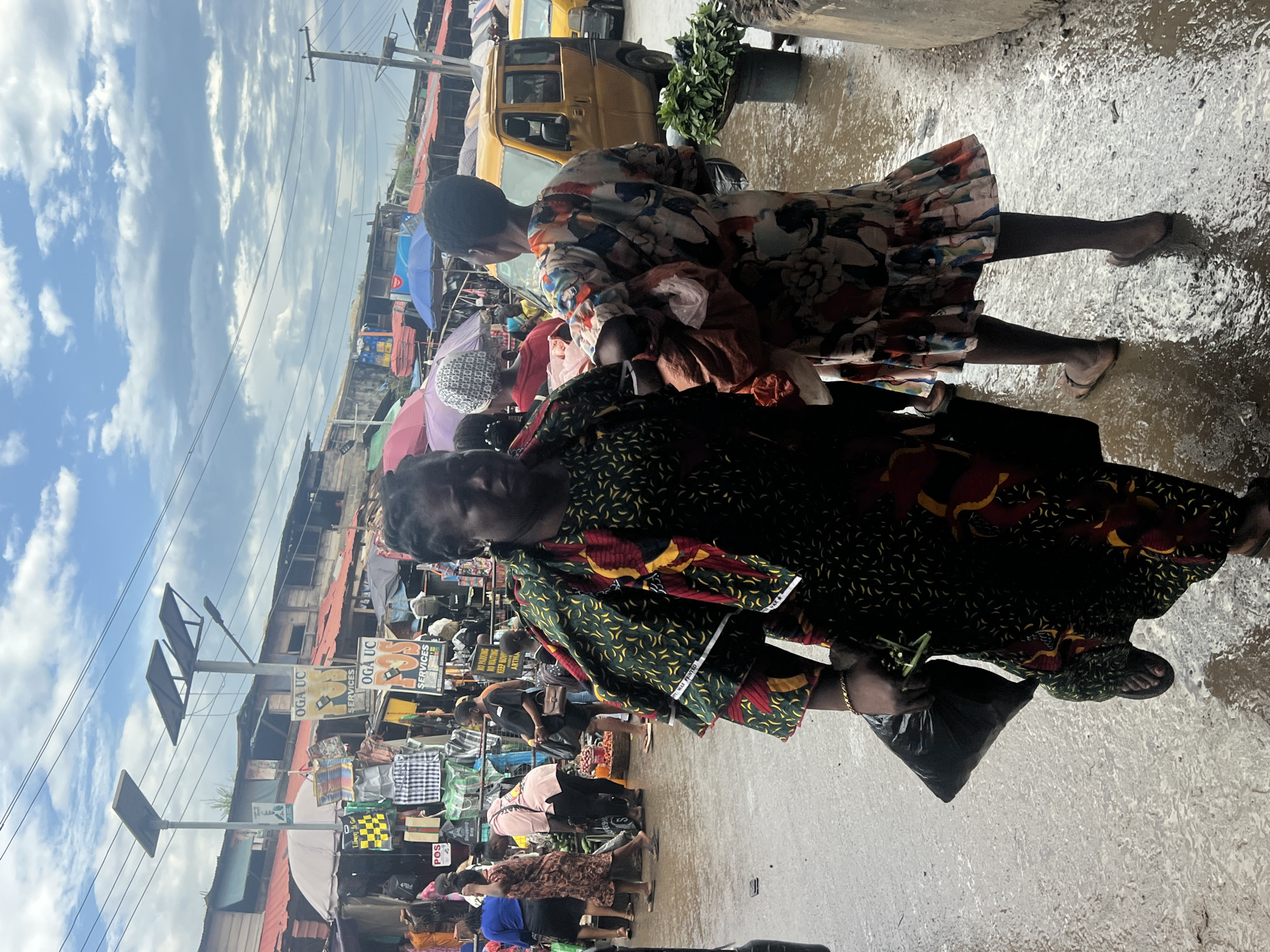 This image captures a bustling market scene in Nkpor, Nigeria, under a cloudy sky. The ground is wet, indicating recent rain, with puddles reflecting the surroundings. In the foreground, two women are prominent. The woman on the left wears a dark green and yellow patterned dress with red accents and carries a black plastic bag. The woman on the right, partially visible, wears a colorful floral-patterned dress and appears to be holding a baby wrapped in a reddish-orange cloth.
Behind them, the market is filled with stalls displaying various goods, including clothing and bags, under umbrellas and awnings. Several other individuals are seen navigating the crowded pathways. Yellow minibusses, typical of Nigerian public transport, are visible in the background, parked along the market street. Solar-powered streetlights are mounted on poles, suggesting a modern infrastructure overlaying the traditional market environment. The overall scene depicts a vibrant and active marketplace in a Nigerian city.