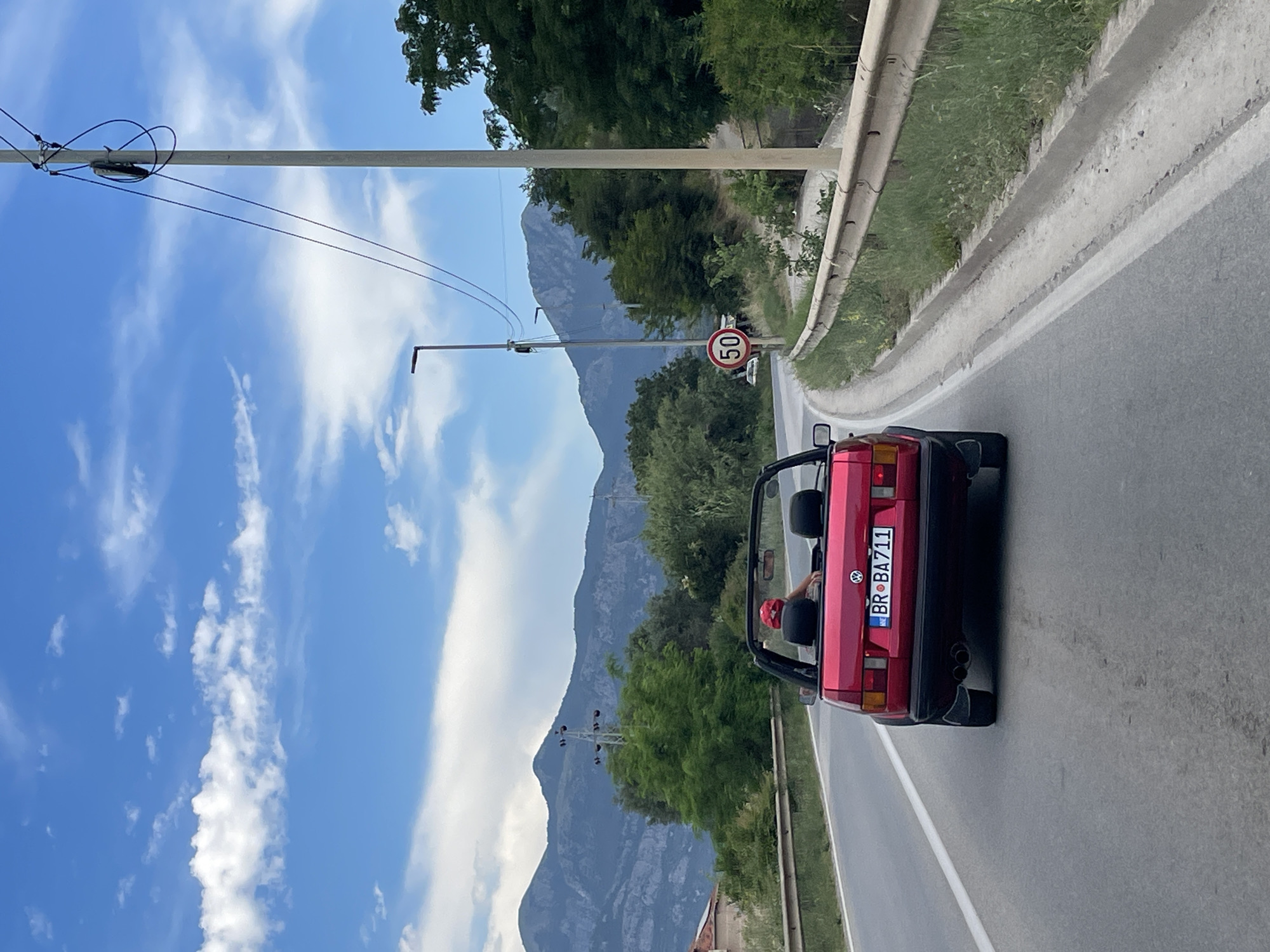 In this roadside scene from Tomba, Montenegro, a red convertible Volkswagen Golf is captured from behind, driving on an asphalt road. The car's license plate reads "BR BA711" with a circular sticker to its right. A person, wearing a red helmet, is visible in the driver's seat, their arm resting on the side of the car. The road is bordered by a metal guardrail on the right, and lush green trees and vegetation line the other side. In the distance, majestic mountains rise against a bright blue sky dotted with fluffy white clouds. A traffic sign indicating a speed limit of 50 is visible on a pole near the roadside. A power line pole with wires stretches across the frame from the left. The image appears to be taken during the day, with clear weather suggesting a pleasant driving experience.