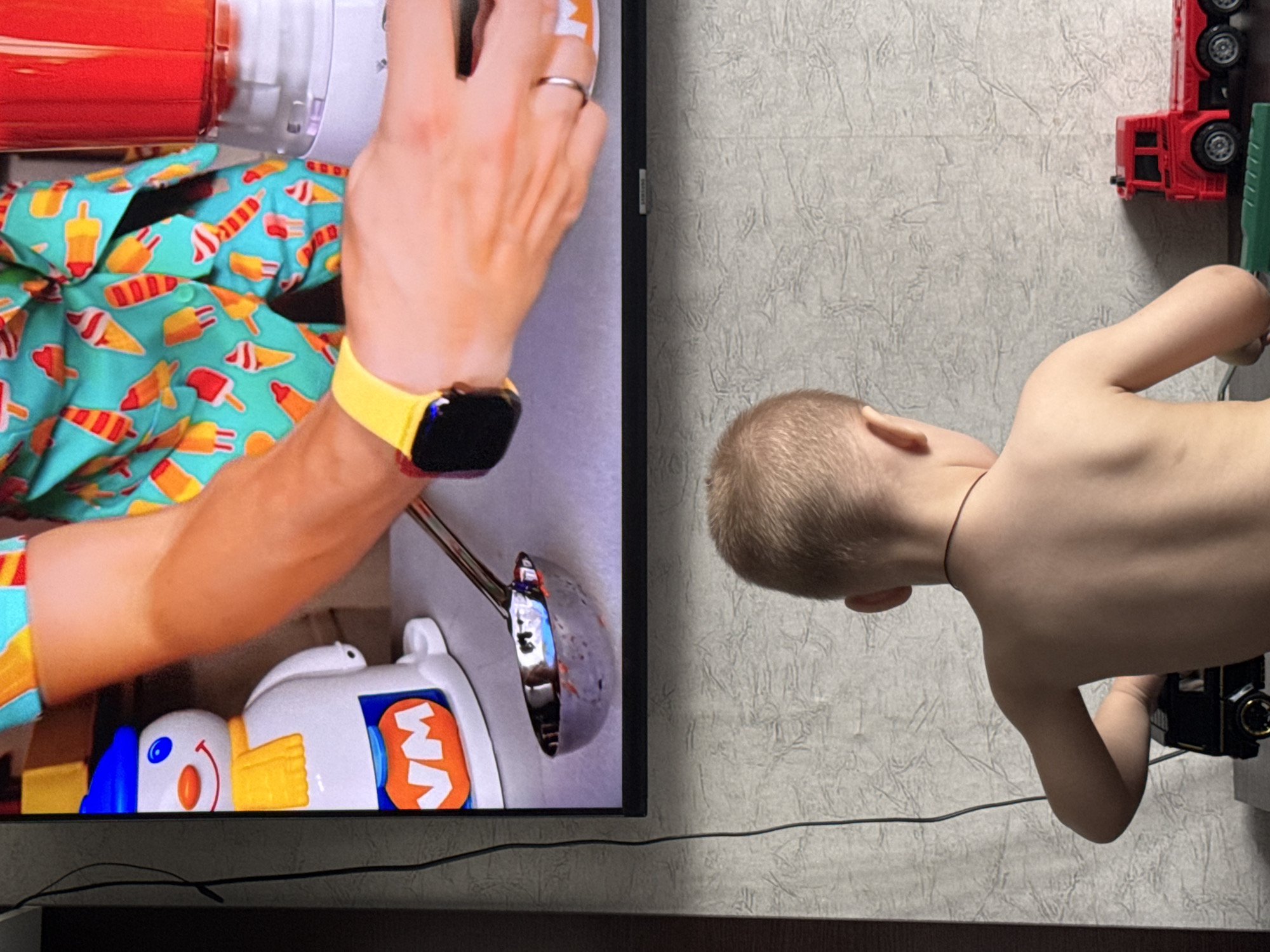 The image shows two subjects: a person's arm and a young child. The arm, adorned with a colorful ice pop patterned shirt and a yellow smartwatch, is positioned over a white toy with a smiling face and blue eyes. The toy also features yellow arms and an orange section with the letters "VM" visible. To the right of the arm, the back of a young child is visible, showing their blonde hair and bare back. The child is leaning towards a toy fire truck, which is red with black wheels and is positioned vertically on a textured, light grey wall. The setting appears to be indoors, with a television screen displaying the arm and toy on the left side of the image. The lighting suggests it might be daytime, though no specific time can be determined. The overall scene appears casual, possibly depicting playtime or a moment of interaction between a child and an adult. The location context is Яромінскі сельскі Савет, Belarus. There is no discernible weather or strong emotional cues.