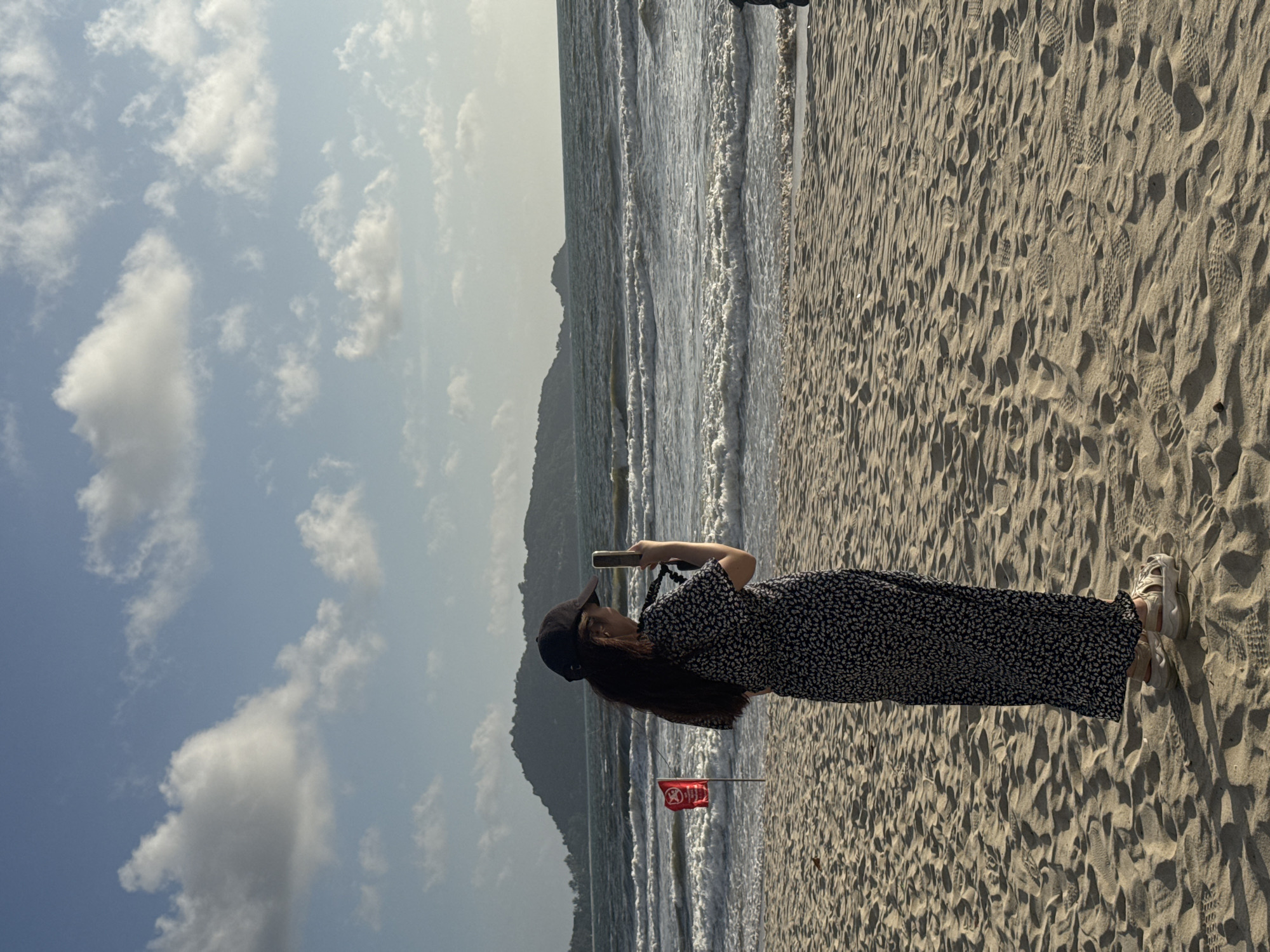 The image shows a person wearing a dark floral print dress and a black baseball cap standing on a sandy beach in Patong, Thailand. The person is facing away from the camera, holding a smartphone in their hands, and appears to be taking a picture or video. In the background, the ocean with waves is visible, and a hilly island can be seen in the distance. A red flag with a white symbol is planted in the sand to the left of the person. The sky is partly cloudy with a clear blue sky peeking through. The sand is textured with footprints.