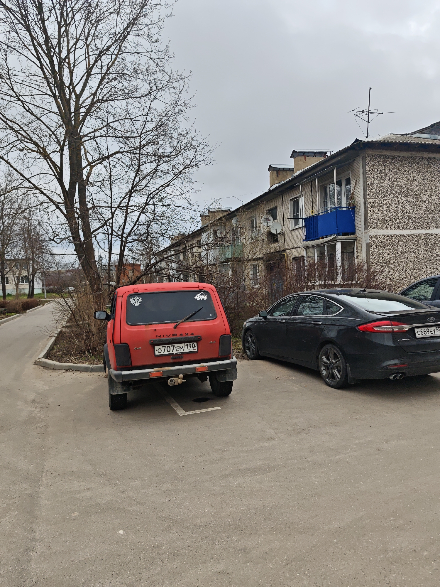 The photograph captures a street scene in посёлок совхоза "Буденновец", Russia, on a cloudy day. In the foreground, a red Niva 4x4 SUV is parked. Behind it, a black sedan is also parked. In the background, a multi-story building with brick and stucco walls is visible. Several satellite dishes are affixed to the building. A bare tree stands beside the red SUV. A narrow sidewalk runs along the left side of the frame.