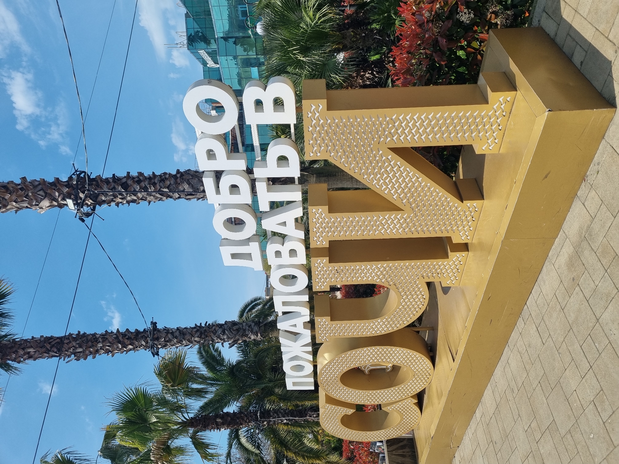 This media file captures a large, welcoming sign in a bright outdoor setting, identified as Sochi, Russia. The main subject is a prominent, three-dimensional sign in Russian, which translates to "Welcome to Sochi." The first part, "ДОБРО ПОЖАЛОВАТЬ В" ("Welcome to"), is rendered in tall, white block letters. The second part, "СОЧИ" ("Sochi"), is displayed in equally large, golden block letters that feature an intricate, textured pattern, possibly representing flying birds or a geometric design, on their visible surfaces.
The sign is situated on a light-colored, paved surface, resembling cobblestones or concrete pavers. In the background, several tall palm trees with visible, textured trunks and vibrant green fronds stand under a clear, bright blue sky with a few scattered white clouds. Overhead, dark utility wires or cables crisscross the frame, passing near the palm trees. Behind the sign and some of the foliage, a modern building with large reflective glass windows is visible, reflecting the sky and surrounding elements. Lush green bushes and some plants with distinctive reddish foliage add depth and color to the backdrop.
The scene depicts a sunny daytime, suggesting pleasant weather. The overall impression is that of a popular tourist destination, emphasizing a warm welcome to the city of Sochi. No people are visible in the image.