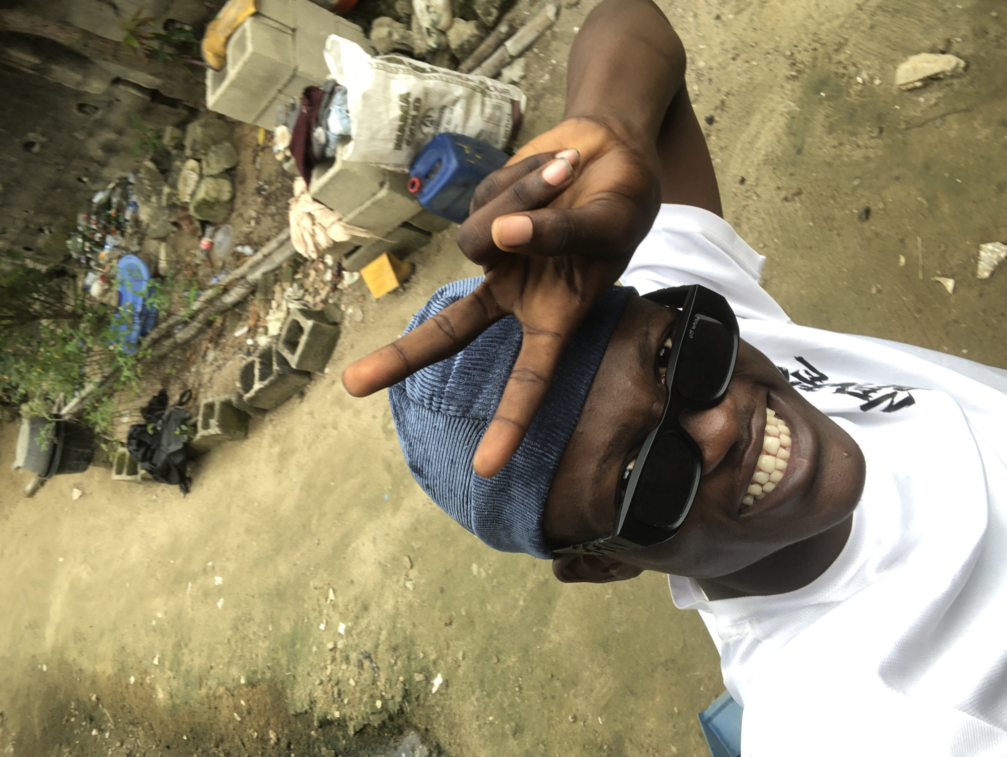 A medium-shot, high-angle selfie is taken outdoors in Ado, Nigeria. A young Black man wearing a blue beanie, black sunglasses, and a white t-shirt smiles broadly at the camera, making a peace sign with his right hand. He is positioned in the foreground, with a dusty, uneven ground and scattered debris filling the background. The debris includes concrete blocks, plastic bags, bottles, and a large blue plastic container. A large white bag with writing is visible in the background, as well as some vegetation and a wall. The overall scene suggests an urban or semi-urban environment with signs of disarray and neglect.