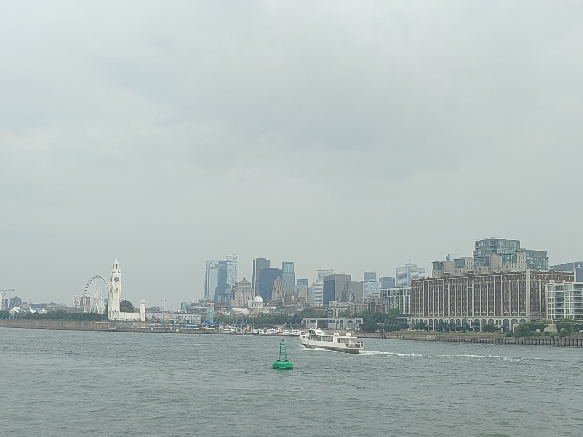 The image shows a wide shot of the Montreal skyline from across the St. Lawrence River on a cloudy day. In the foreground, the choppy, grey water of the river is visible. A small, white boat with a dark roof is moving from right to left, leaving a wake. A green buoy with a white top is positioned in the water to the left of the boat.
Along the riverbank, a line of buildings forms the city's edge. The most prominent structures on the left side of the skyline include the large white Ferris wheel of La Grande Roue de Montréal and the adjacent clock tower. To the right, a dense cluster of modern skyscrapers and older brick buildings rises, creating a varied urban landscape. A large, multi-story brick building with arched windows is particularly noticeable on the far right. Trees and greenery line the riverbank in front of the buildings. The sky is overcast and a uniform light grey, obscuring any distinct cloud formations. No people are clearly identifiable in the image, though there appear to be a few figures on the deck of the moving boat.
