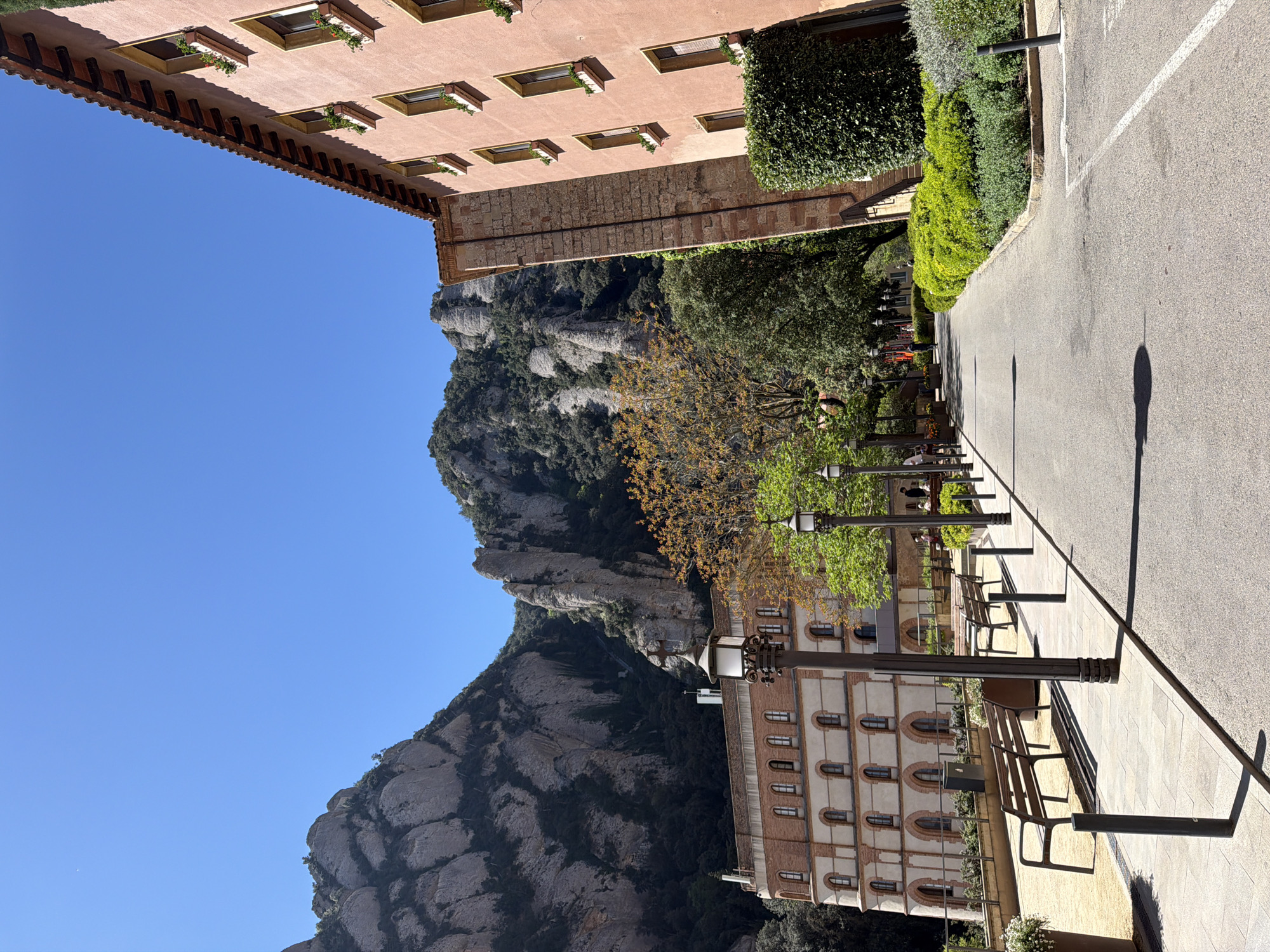 The image captures a sunlit outdoor scene at Monistrol de Montserrat, Spain, featuring a striking blend of natural beauty and architecture. In the background, the iconic, rugged peaks of the Montserrat mountain range dominate, characterized by their unique grey, rocky formations and patches of dark green vegetation, all set against a clear, brilliant blue sky.
Two distinct multi-story buildings frame the scene. On the left, a reddish-orange building with a tiled roof features several windows, each equipped with a planter box containing small green plants. On the right, a lighter-colored building, possibly beige or white, exhibits a series of arched windows with red brick detailing.
A wide, paved pathway or road extends through the center of the image, appearing to be a pedestrian area or courtyard. This path is lined with several dark streetlights and benches. A few indistinct figures can be seen sitting on the benches or walking in the distance, suggesting leisurely activity. Lush green hedges and trees, some with sparse, yellowish-orange leaves, border the path and contribute to the vibrant foreground. Strong shadows cast by the streetlights and buildings indicate bright daylight, suggesting the photograph was taken on a clear, sunny day, likely mid-morning or afternoon. The scene conveys a sense of grandeur and tranquility, with the imposing mountains providing a magnificent backdrop to the well-maintained surroundings. No discernible text is visible in the image.