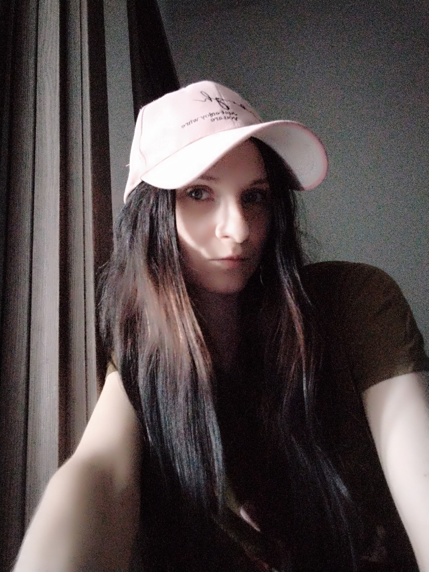 A young woman with long, dark hair and a light pink baseball cap is photographed from the chest up. She is wearing a dark olive green t-shirt. The background consists of vertical dark gray curtains on the left and a textured gray wall on the right. The lighting casts a shadow across the right side of her face, illuminating her left eye and cheek. The cap has white lettering that is difficult to read. The image appears to be a selfie, as her arm and a portion of her hand are visible on the left side of the frame.