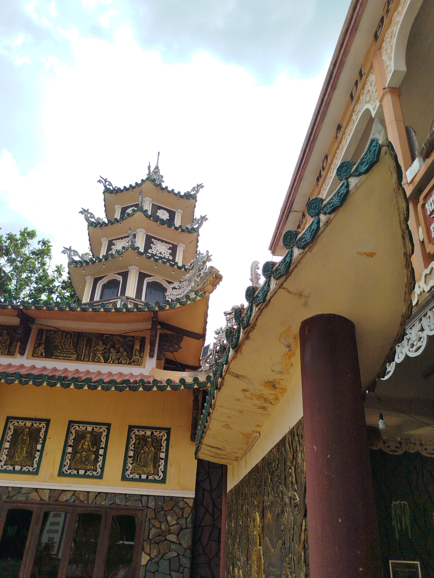 A low-angle shot captures a yellow three-tiered pagoda-style building and the corner of a building with a column in Phú An, Vietnam. The pagoda has a tiered, ornamented roof with green detailing and decorative elements. Below the roof, there are panels depicting figures. The lower section of the pagoda has three framed pictures of seated figures above a stone wall and windows. The other building has a curved, green-detailed roof supported by a round, brown column, and decorative trim. The sky above is light with scattered clouds.