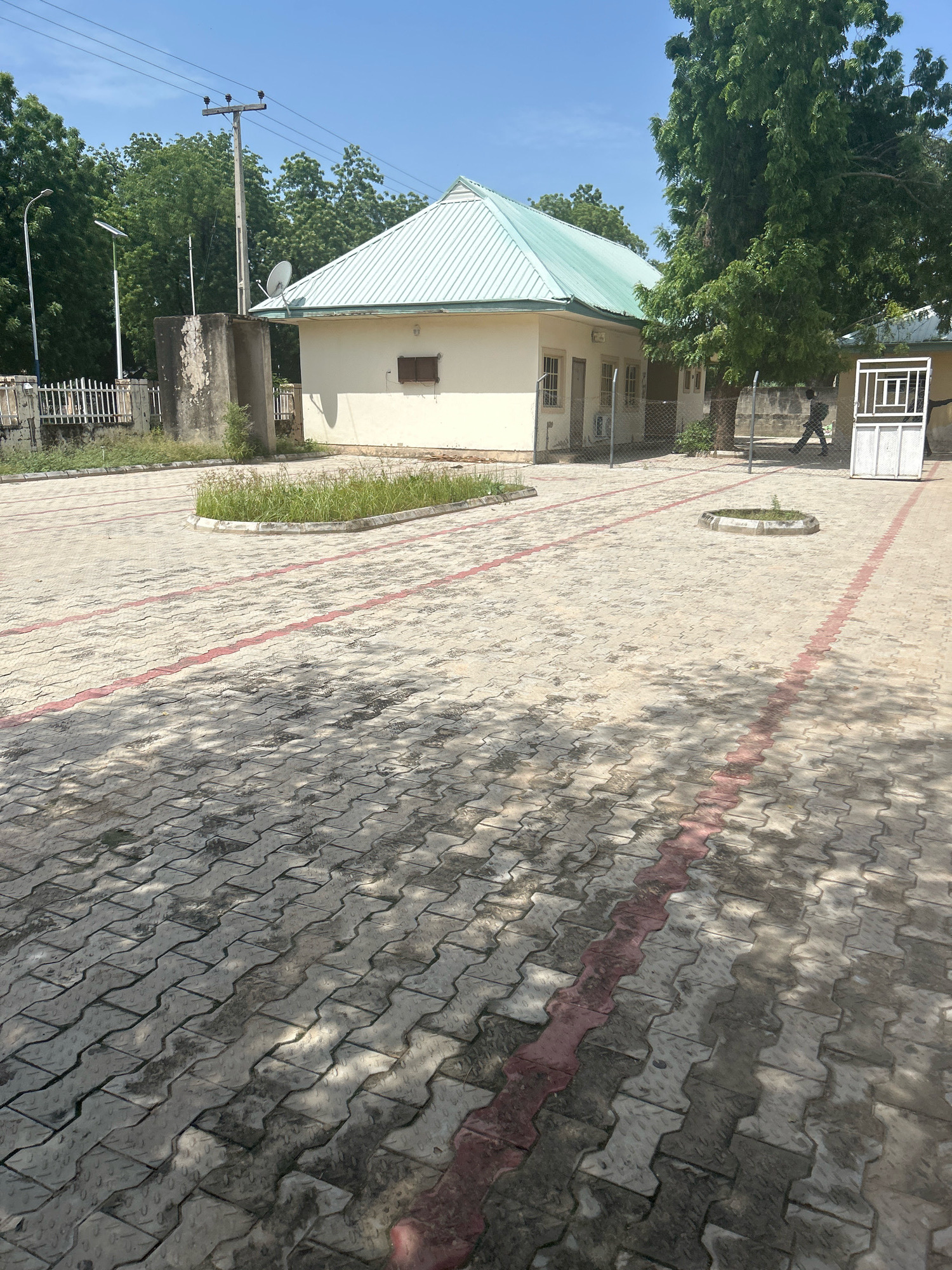 The image depicts an outdoor scene in Mairi, Nigeria. In the foreground, a paved area with interlocking concrete blocks, some with red accents, suggests a courtyard or walkway. The paving is partially shaded by trees. In the mid-ground, a light-colored building with a green metal roof is visible. The building has several windows and a small satellite dish is mounted on its side. To the left of the building, a concrete structure with a power pole and electrical wires stands. Trees and foliage are present in the background. On the right side of the image, a white metal gate is visible, and a person is walking away from it, towards the building. The overall environment appears to be a moderately maintained public or institutional space.