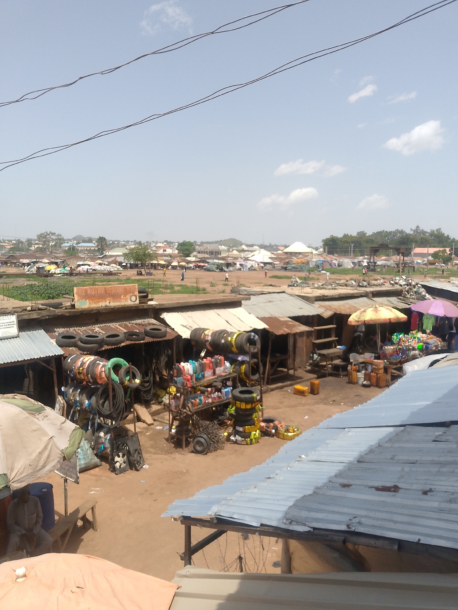 The image shows an open-air market in Bukuru, Nigeria. Predominantly visible are metal-roofed stalls offering automotive parts like tires, engine oil, and assorted vehicle components. The ground is dusty, and the stalls are clustered together. Further in the background, there's a wider, open area with more stalls and people moving around. In the distance, there is a blurry cityscape. Overhead, electrical wires cross the sky, which is partly cloudy. A person is sitting on a bench near the front left, under an umbrella.