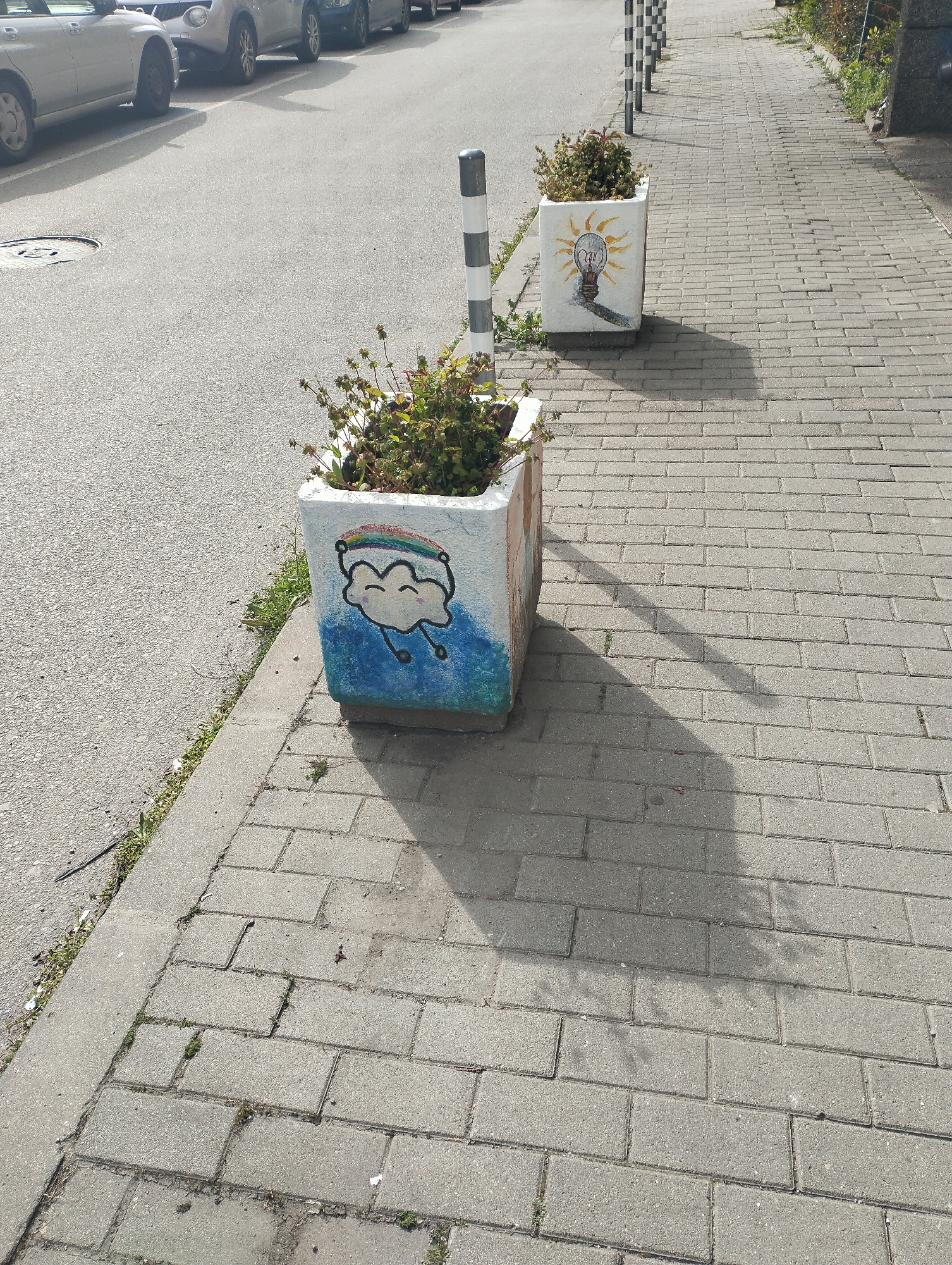 The image shows two square, painted concrete planters with vegetation in them. They are located on a brick paved sidewalk next to an asphalt road. The first planter features a cartoon-like cloud with a rainbow overhead and blue raindrops, while the second planter shows a light bulb drawing. The planters are spaced a few feet apart, and a row of black and white striped bollards are in the background. The scene is lit by sunlight. Based on the bollards and street features, the location is likely in Sofia, Bulgaria. Cars are parked along the side of the road. There are no people visible in the image.