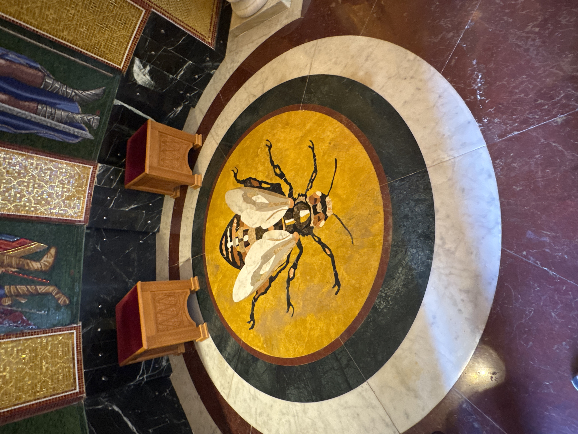 The image shows a floor mosaic of a bee. The mosaic is circular and features the bee in the center on a yellow background, surrounded by concentric circles of dark green, white, and maroon. The bee is detailed with dark legs, a patterned body, and light-colored wings. The mosaic is situated in an ornate location, possibly a church or other significant building, as indicated by the nearby mosaic art on the floor, gold-colored tile, and decorative wooden stools with red velvet tops. The location is likely Tsakistra, Cyprus, as the bee mosaic is a notable feature there. No people are visible in the shot. The camera is positioned directly above and slightly tilted, which gives a clear view of the bee mosaic and its surroundings.