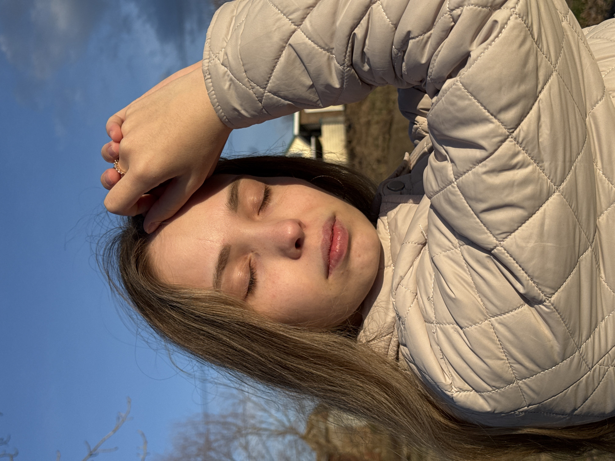 This close-up, portrait-style image, taken in Chaikovsky, Russia, features a young person, likely female, resting outdoors. The subject has long, light brown hair and her eyes are closed, suggesting a moment of peace, relaxation, or weariness. Her left hand, adorned with a delicate, intricately designed ring on the ring finger, is placed over her forehead, possibly shielding her eyes from the sun or indicating a thoughtful state.
She is wearing a light beige or cream-colored quilted jacket, which shows clear diamond-patterned stitching and soft folds. The warm, directional sunlight highlights the textures of her hair and jacket, and casts gentle shadows on her face.
The scene is set against a clear, bright blue sky, with hints of blurred background elements that resemble bare tree branches and a distant structure, suggesting an outdoor environment during a season without lush foliage, possibly late autumn or early spring. The bright sky and visible sunlight indicate it is daytime. No visible text is present in the image.