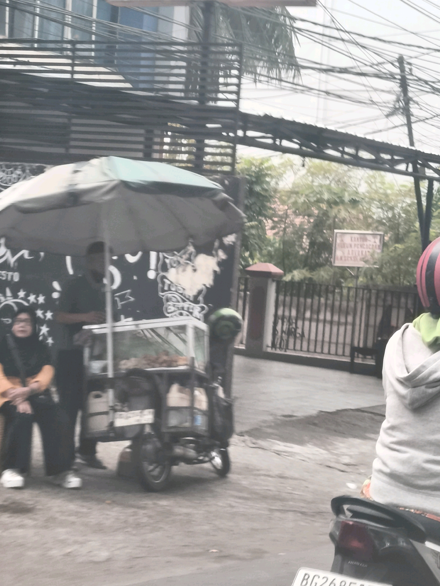 An exterior medium shot depicts a street scene in Palembang, Indonesia, on an overcast day. In the foreground, a person in a light-colored hoodie and black helmet is seated on a motorcycle, facing away from the camera. The motorcycle's rear is visible, including a license plate that reads "BG2685F". To the left, a food cart with a glass display case is parked under a green umbrella. A vendor, a person wearing a dark shirt and pants, stands behind the cart. A person wearing a headscarf and an orange top is seated to the left of the food cart. The background shows a building with horizontal slatted awnings, trees, a fence, and a sign with Indonesian text. The ground is wet, indicating recent rain.