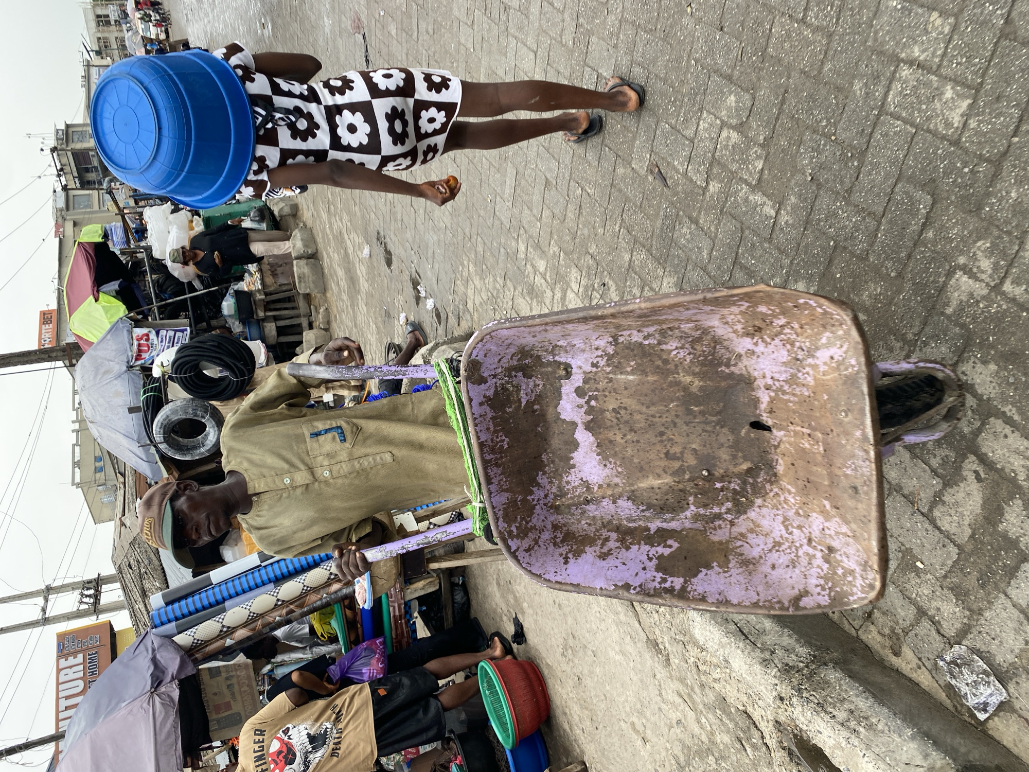 A street scene in Lekki, Nigeria, shows a man standing beside a wheelbarrow. He wears a tan shirt, pants, and a cap. The wheelbarrow's basin is weathered with remnants of purple paint. Rolls of patterned linoleum and electrical wires are stacked behind him. Nearby, a person is carrying a blue plastic tub on their head and wearing a black and white dress. The pavement is paved with light grey bricks. A variety of businesses are visible, marked with signs, tents, and umbrellas. Another individual wearing a tan t-shirt and black shorts is present in the scene.