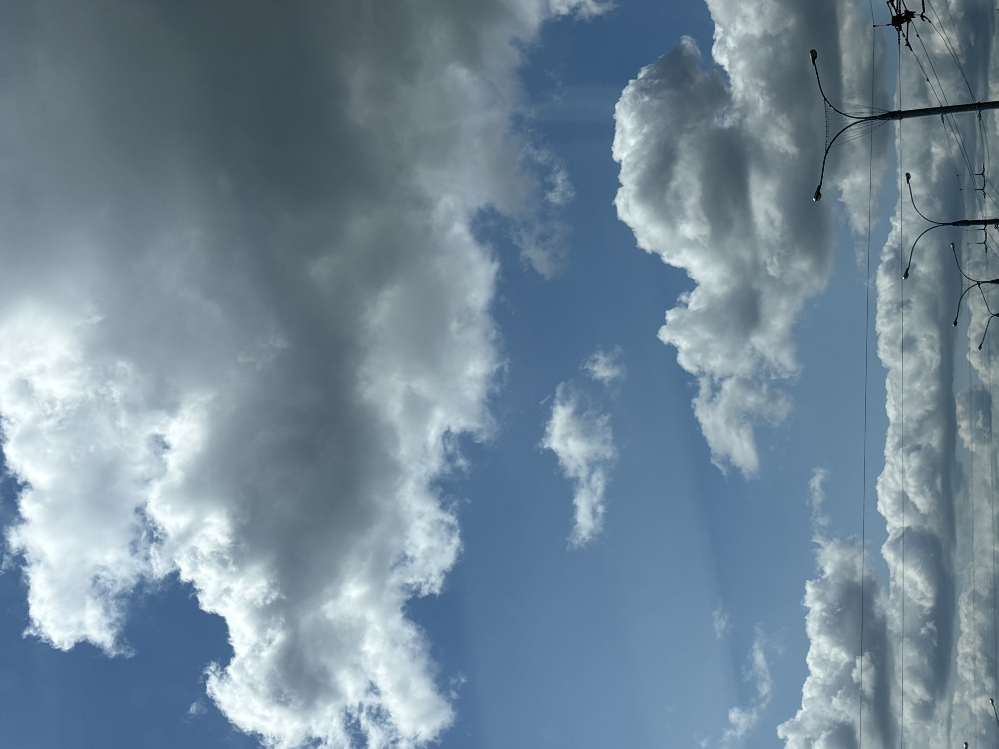 The image shows a bright blue sky with scattered clouds, some of which are dark and ominous, while others are bright white. The clouds appear to be cumulus clouds. The scene is framed by utility poles and wires running horizontally across the lower part of the image, suggesting an urban or suburban environment. Some streetlights are also visible. The lighting suggests it is daytime, with the sun likely positioned above and to the left of the frame, casting shadows on the clouds and illuminating the sky. There are no people or vehicles visible in the image. Based on the presence of streetlights and utility infrastructure, and the style of the streetlights which are common in Eastern Europe, this scene could be in Minsk, Belarus.