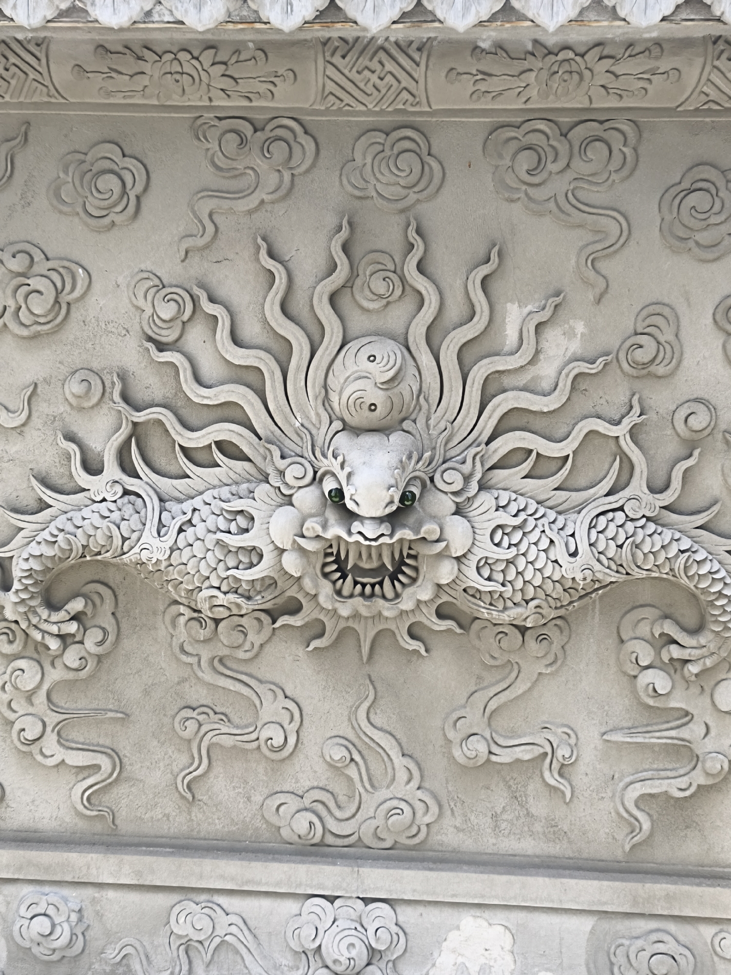 The image is a close-up, eye-level, full shot of a bas-relief carving of a dragon. The dragon is depicted in a stylized, serpentine manner, with its body extending horizontally on either side of its head. The carving is monochromatic, appearing to be made of stone or concrete, with a light grey hue. The dragon's head is central, facing forward, with a fierce expression. It has prominent eyes with green irises, sharp teeth bared in a snarl, and flowing mane-like tendrils radiating from its head. Scales cover its body, and it is surrounded by swirling cloud motifs, a common element in East Asian art. The carving is detailed, with clear definition between the scales, teeth, and tendrils. The top of the image shows a decorative border with geometric patterns and floral motifs. The bottom of the image reveals a lower section of the wall, also adorned with carvings. The overall impression is one of power and intricate artistry. The location is listed as Unknown, Unknown.