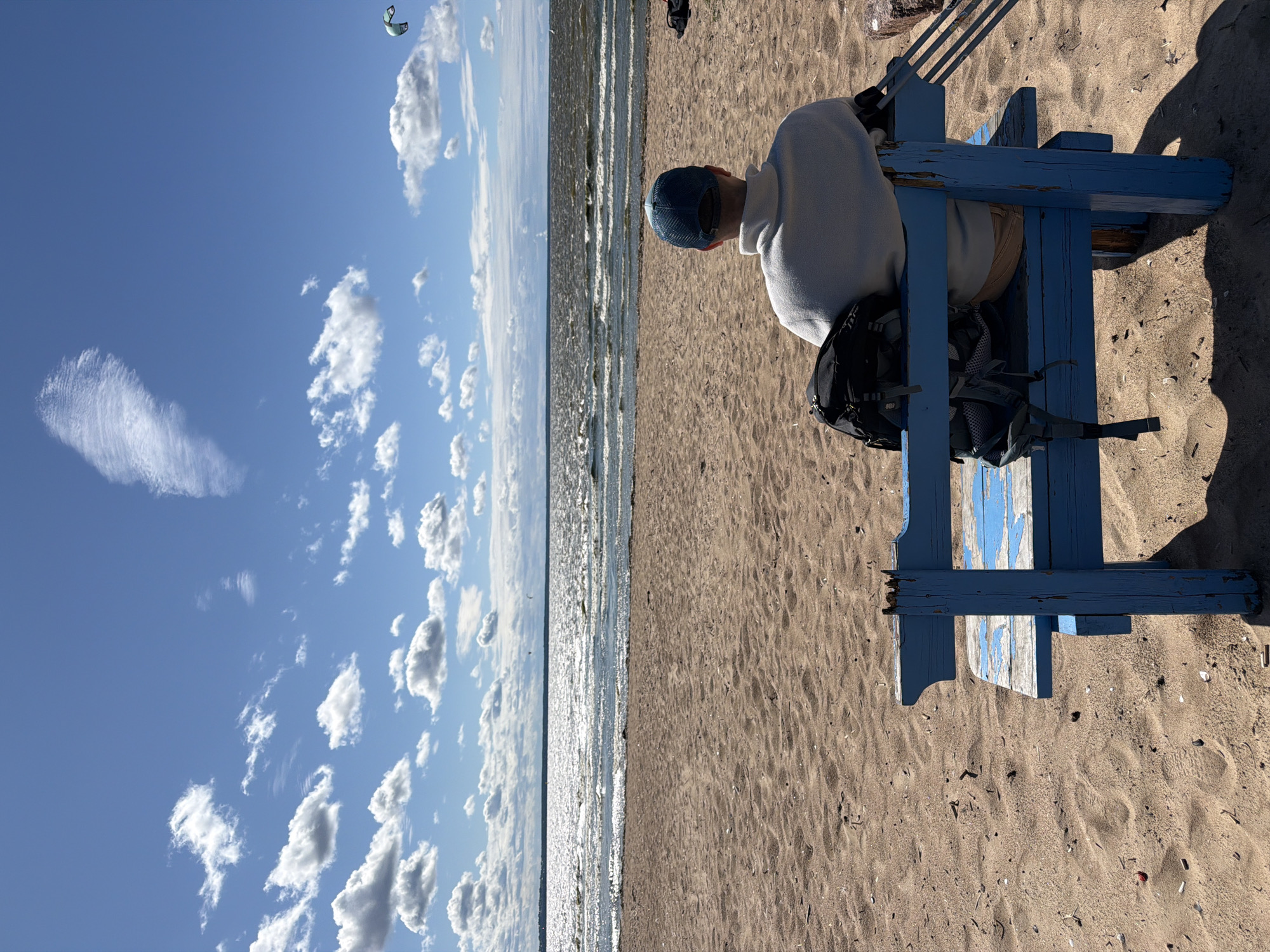 The image depicts a sunny day on a sandy beach, likely in Kronstadt, Russia, with a person sitting on a weathered blue wooden bench facing the water. The sky is a vibrant blue with scattered white clouds, and a faint, wispy cloud stretches across the upper portion of the frame. The sea is choppy with sunlight glinting off the waves, and a distant shoreline is visible. In the sky, a kite surfer is visible as a small speck in the upper right corner. The person on the bench is wearing a light-colored sweatshirt and a blue baseball cap, with a black backpack resting on the bench beside them. The bench itself shows signs of wear, with peeling blue paint. The beach sand is tan and appears relatively clean, with a few small pieces of debris scattered on its surface.