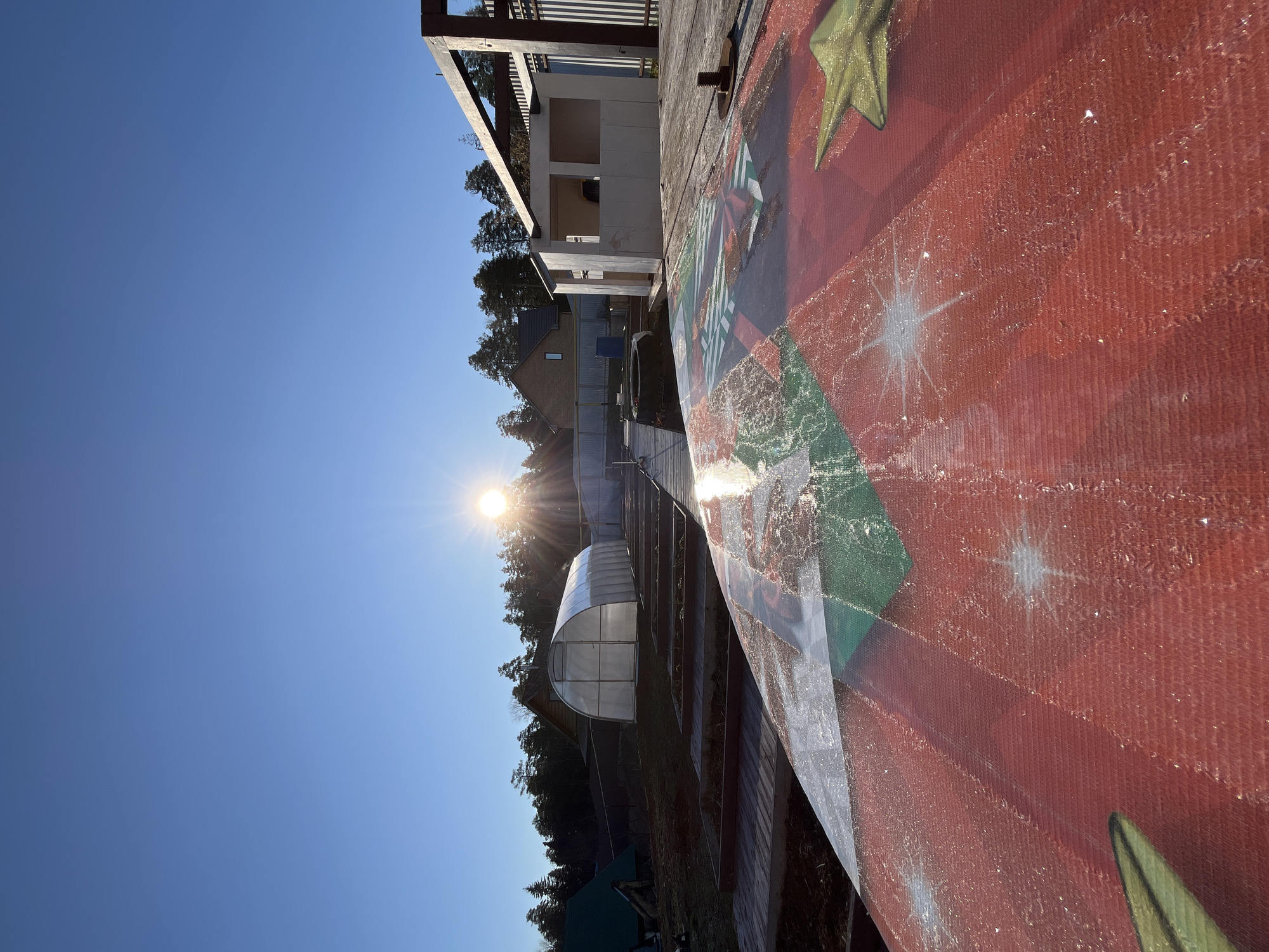 The image captures an outdoor landscape scene, likely taken in Chaikovsky, Russia, under clear, sunny conditions. The perspective is tilted, showing a broad vista from what appears to be an elevated position, possibly a deck or similar structure.
In the immediate foreground, a large, textured surface occupies the right side of the frame. This surface, possibly a boat hull or a decorative panel, is painted with vibrant red, green, and white patterns, including golden stars and what appears to be glitter or sparkling elements. A prominent metal bolt is visible on an adjacent wooden plank.
Moving towards the mid-ground, a pathway or open ground leads to several structures. These include a multi-story building with an exposed wooden frame on top, a house with a dark roof and lighter walls, and a distinctive semi-circular, translucent greenhouse. Behind these buildings, a dense line of evergreen trees forms a natural barrier, suggesting a forest.
The sky is a clear, bright blue, devoid of clouds. The sun is a dominant feature, positioned low on the left side of the frame, emitting strong rays and creating a sunburst effect and noticeable lens flare. This light suggests either early morning or late afternoon.
No people or specific activities are visible, presenting a static and serene view. The bright sunlight and clear sky indicate pleasant weather. No discernible text is visible in the image.