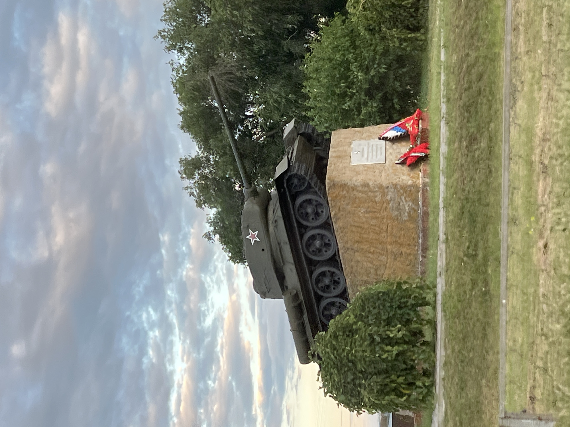 The image shows a Soviet-era T-34 tank monument at the entrance to the city of Zimovniki, Russia. The tank, painted olive drab, is mounted on a concrete pedestal covered with light-colored stone. A red star with a white outline is visible on the side of the tank's turret. In front of the pedestal, there is a plaque and a memorial wreath with red and blue ribbons. The monument is surrounded by green trees and bushes, with a grassy area in the foreground. The sky above is cloudy, with hues of gray and pale yellow, suggesting either dawn or dusk.