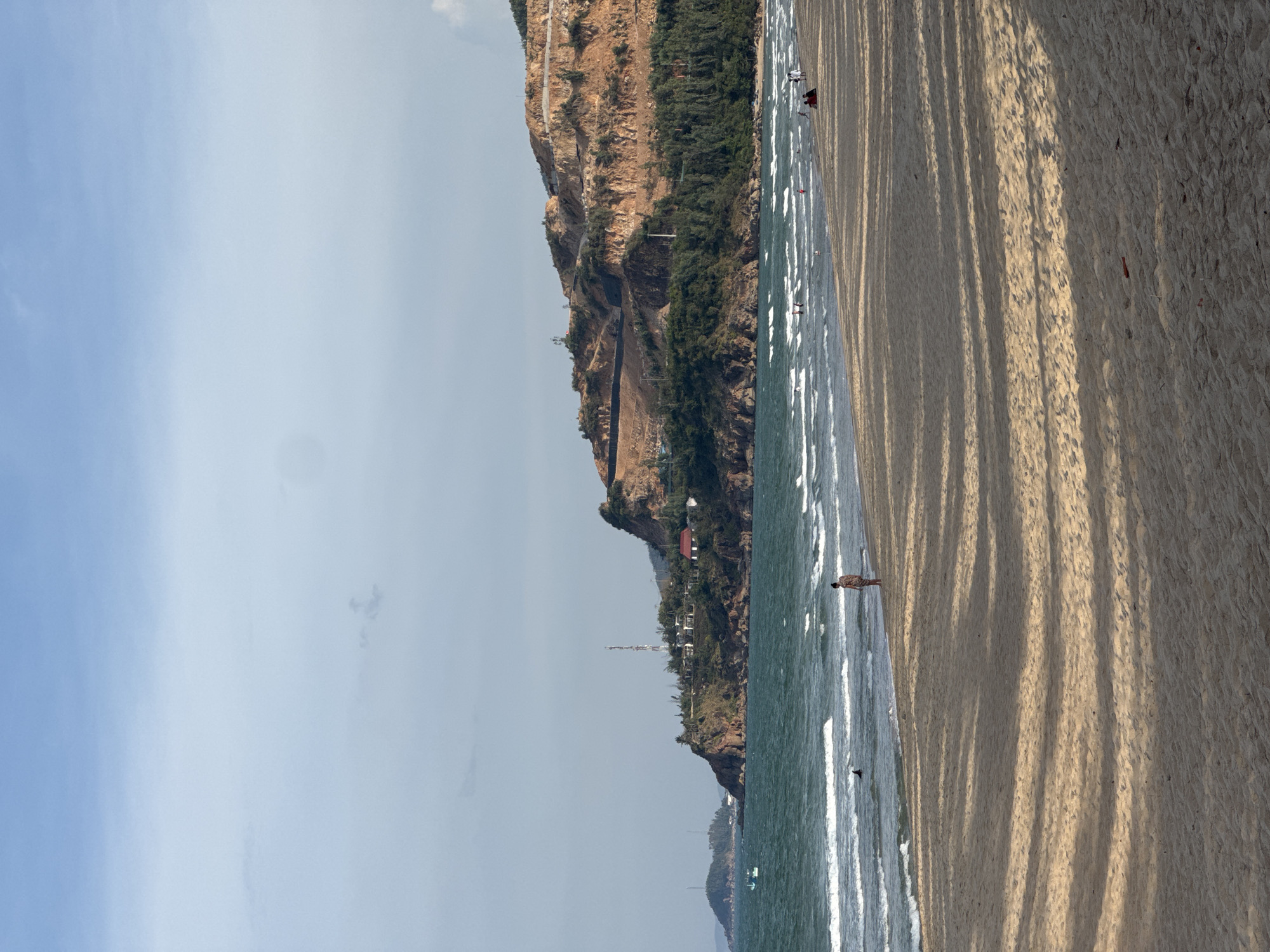 The image depicts a beach scene in South Quy Nhon Ward, Vietnam. The composition is dominated by a sandy beach in the foreground, casting long shadows from an unseen source, suggesting it is late afternoon or early morning. The ocean stretches along the left side of the frame, with gentle waves lapping the shore. In the background, a steep, rocky cliff face rises, covered in patches of green vegetation and dotted with buildings. A road winds its way up the side of the cliff, and a tall communication tower is visible near the crest. A few individuals can be seen on the beach and in the shallow water, engaging in activities like walking or wading. The sky is clear and light blue, indicating fair weather. The overall atmosphere is serene and tranquil, conveying a sense of peacefulness associated with a seaside environment.