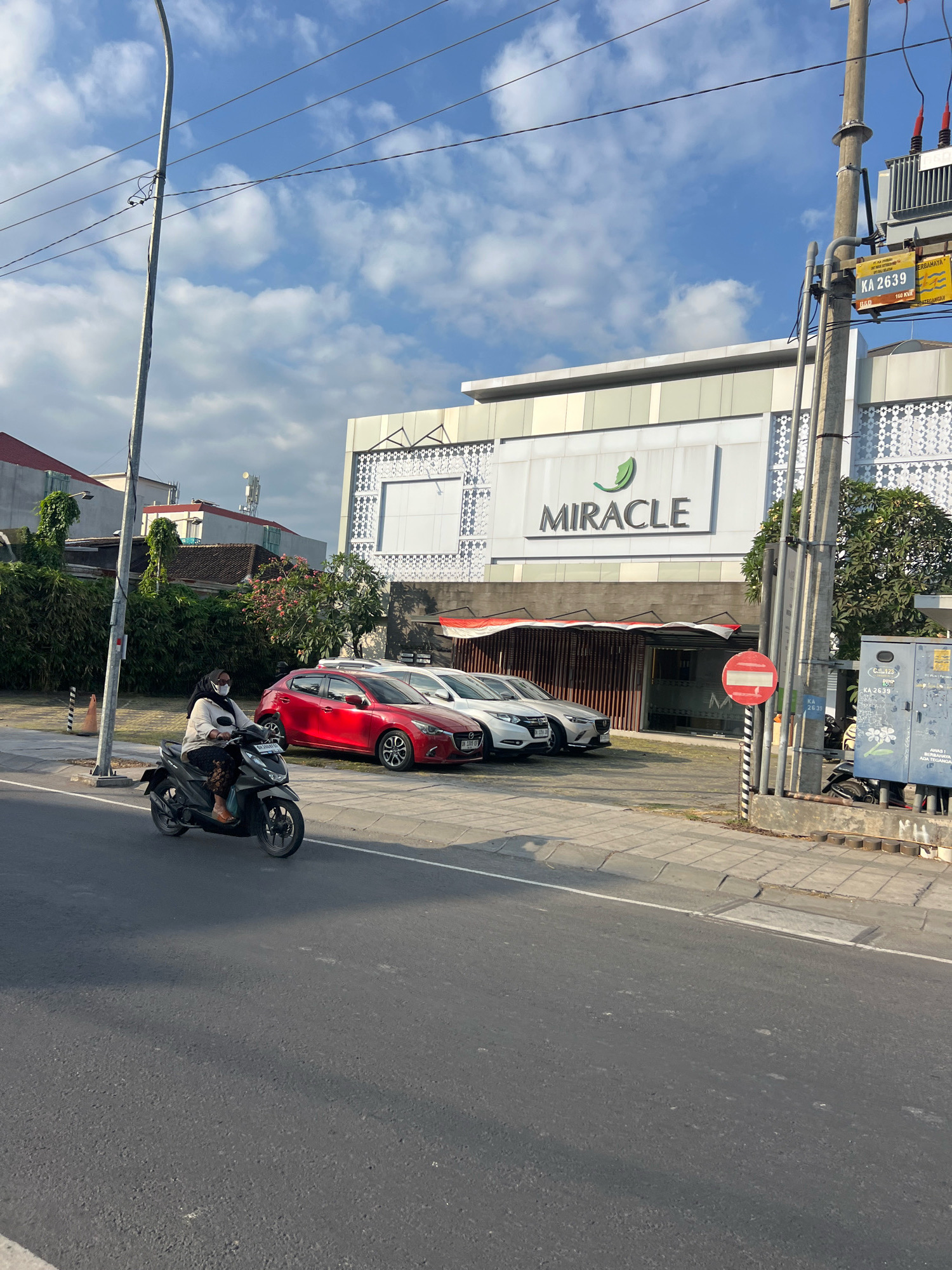 The image depicts a street scene in Kuta, Indonesia. In the foreground, a person wearing a mask and hijab rides a dark-colored scooter on the asphalt road. The rider is wearing a light-colored top and dark patterned pants, with brown sandals. Behind the scooter, parked on the side of the road, is a red car. Further back, several white cars are parked. A building with the prominent signage "MIRACLE" and a green leaf logo is visible. The building has a modern facade with patterned decorative elements and large glass windows. A traffic light pole and electrical equipment, including a transformer, are also present. The sky is blue with scattered white clouds. Trees and lush greenery are visible on the left side of the image. A "no entry" sign is posted near the electrical pole. The overall scene suggests a commercial or retail area in Kuta.